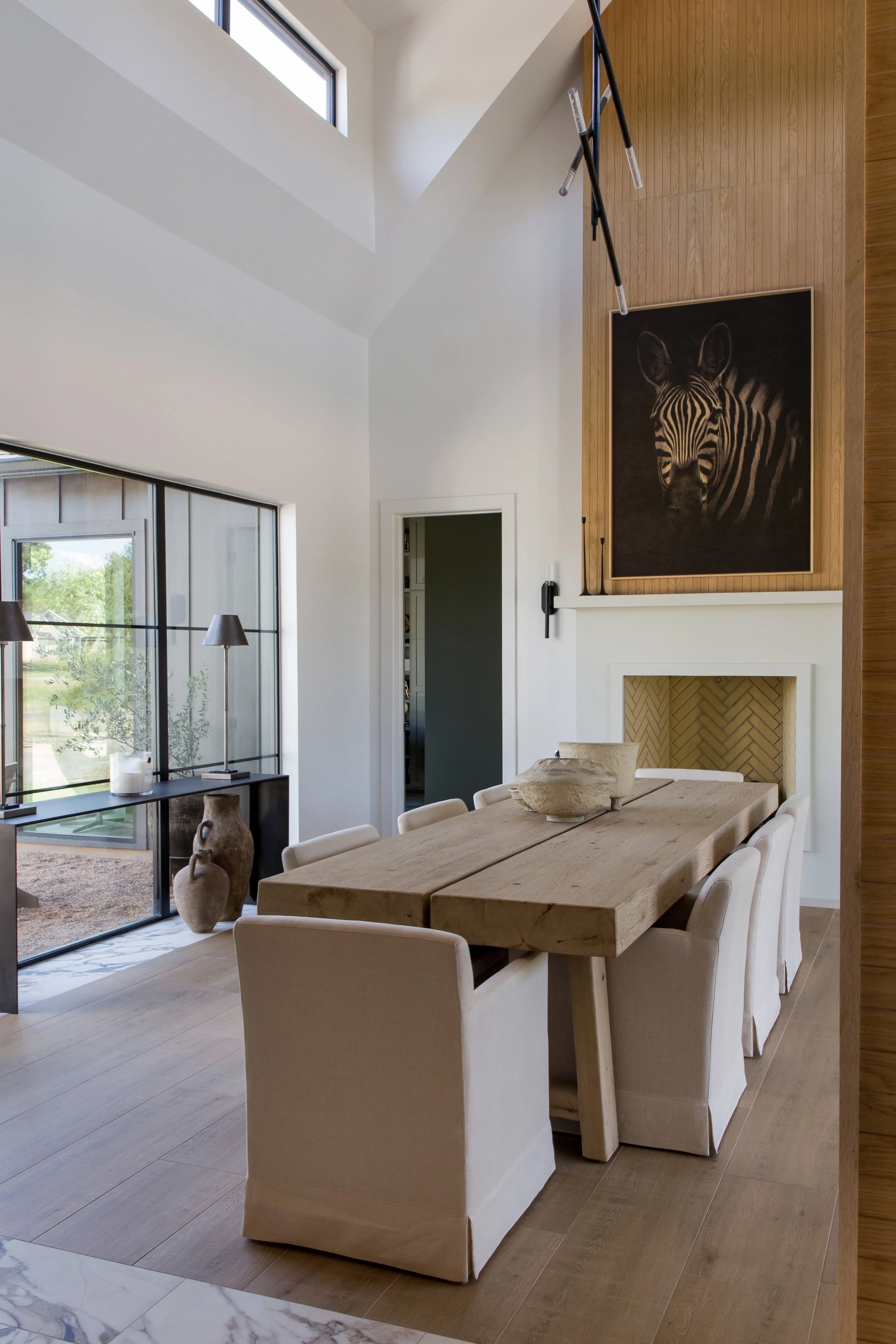 Modern dining room with wooden table, beige dining chairs, large windows, and zebra artwork above a fireplace.
