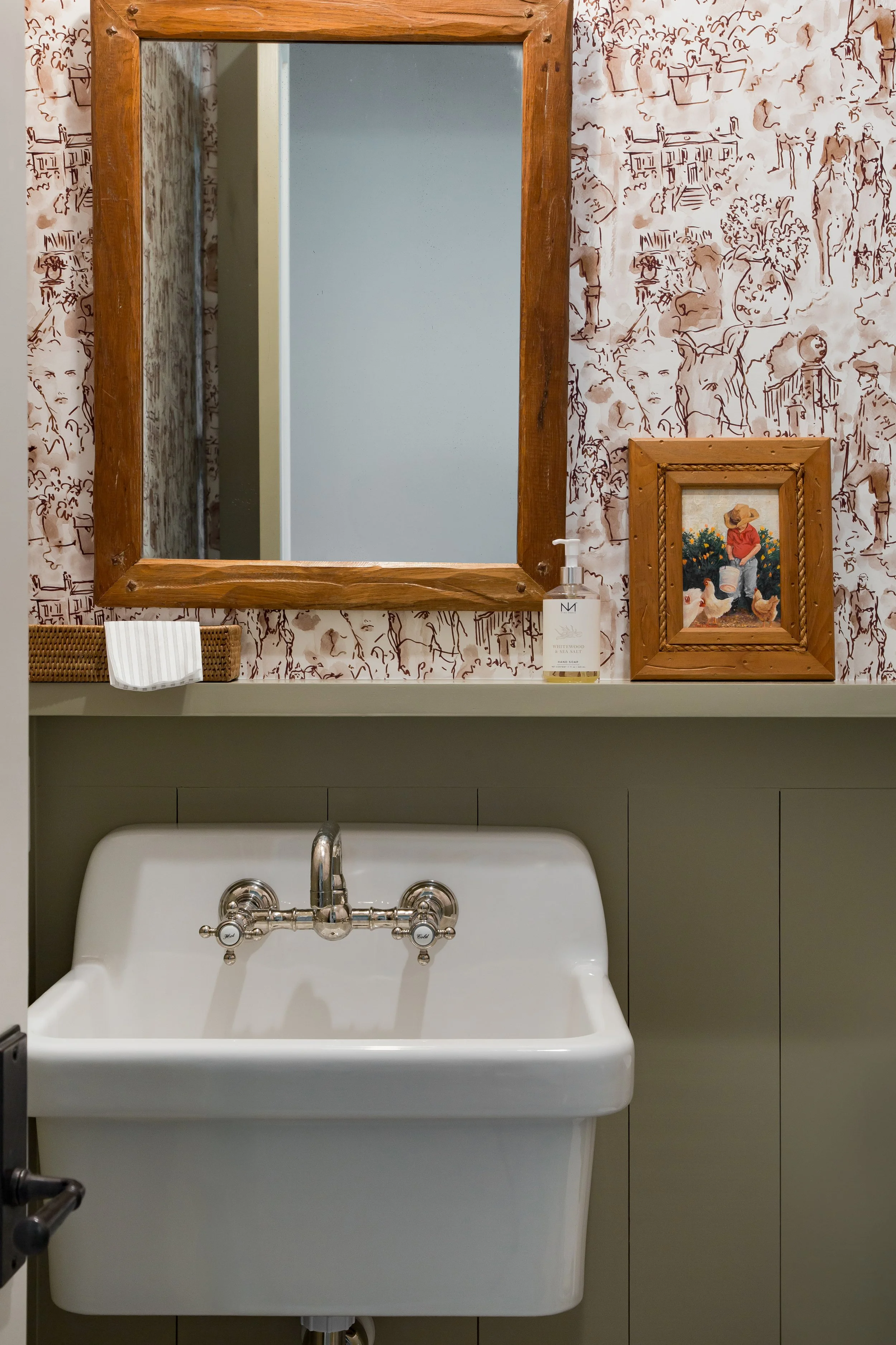 A farmhouse-style bathroom sink with a vintage faucet, a large wooden-framed mirror, a small framed painting of a child with chickens, and a bottle of hand soap on a shelf with wallpaper featuring a sketch-like pattern of people and buildings.