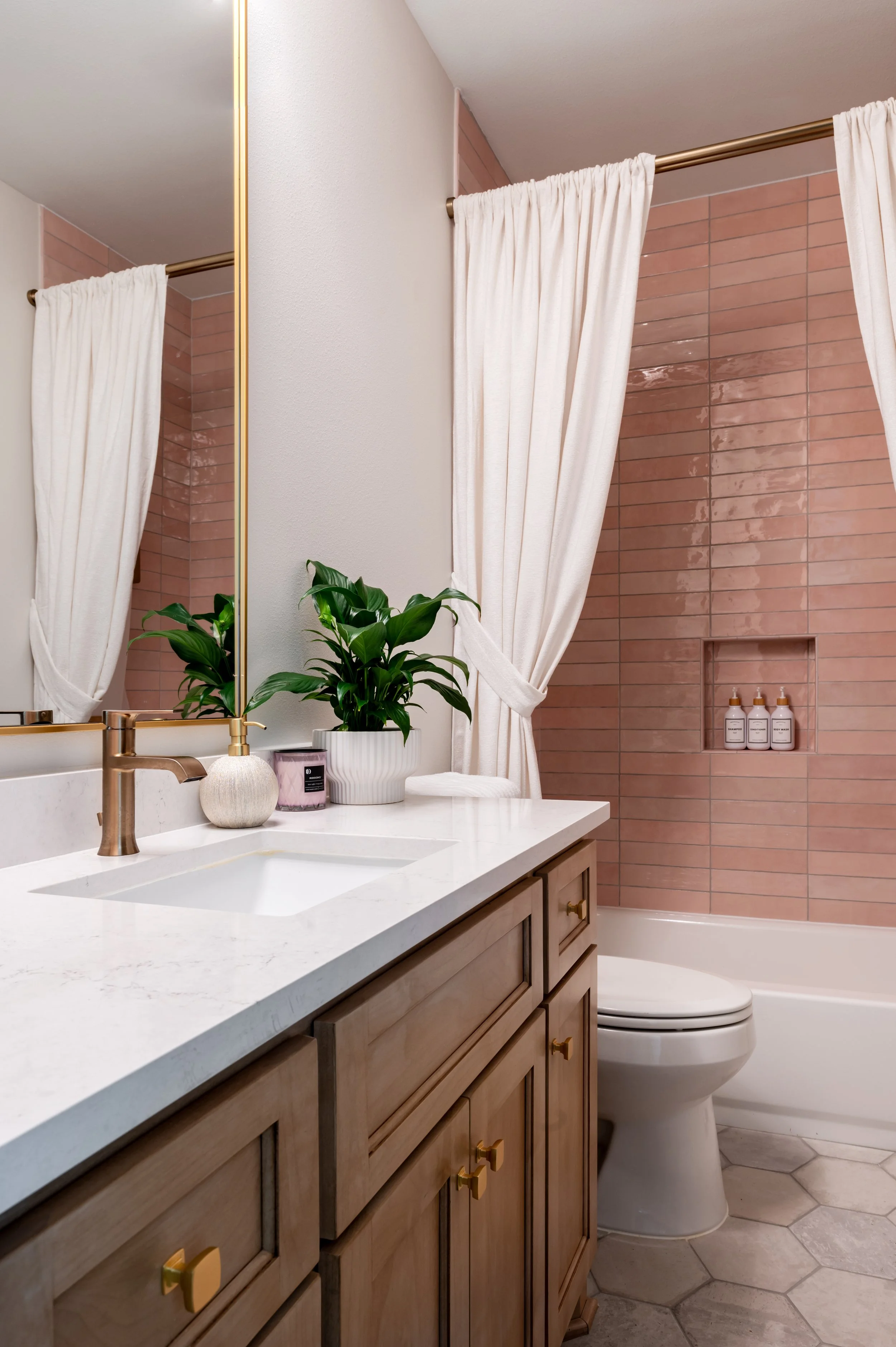 Bathroom with a wooden vanity, white countertop, potted plant, and a large mirror. Shower area has pinkish tiles with built-in shelves containing bottles.