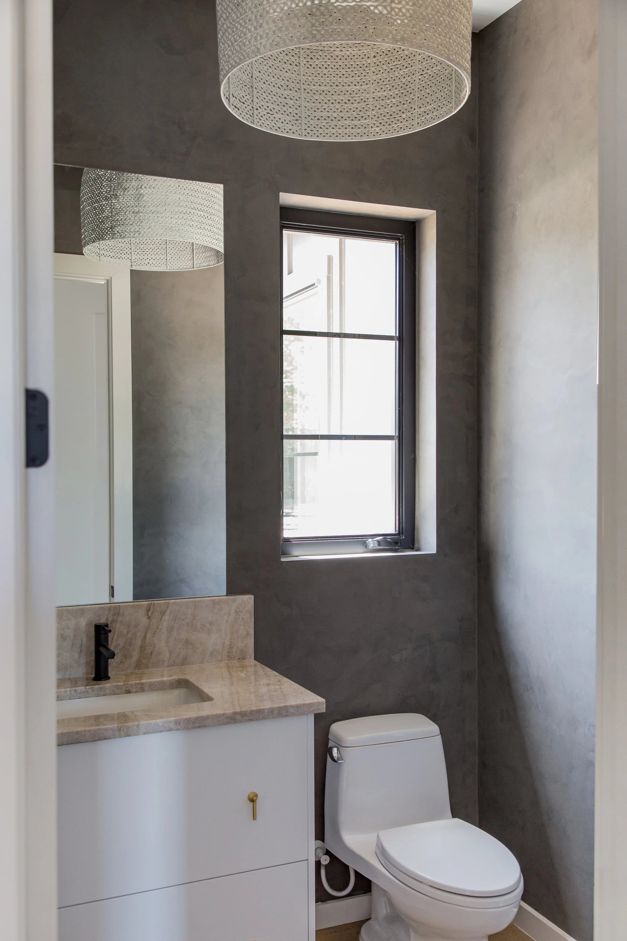 A small bathroom with a dark gray textured wall, a window, a white toilet, and a beige marble countertop with a sink. Two white pendant lights hang from the ceiling.