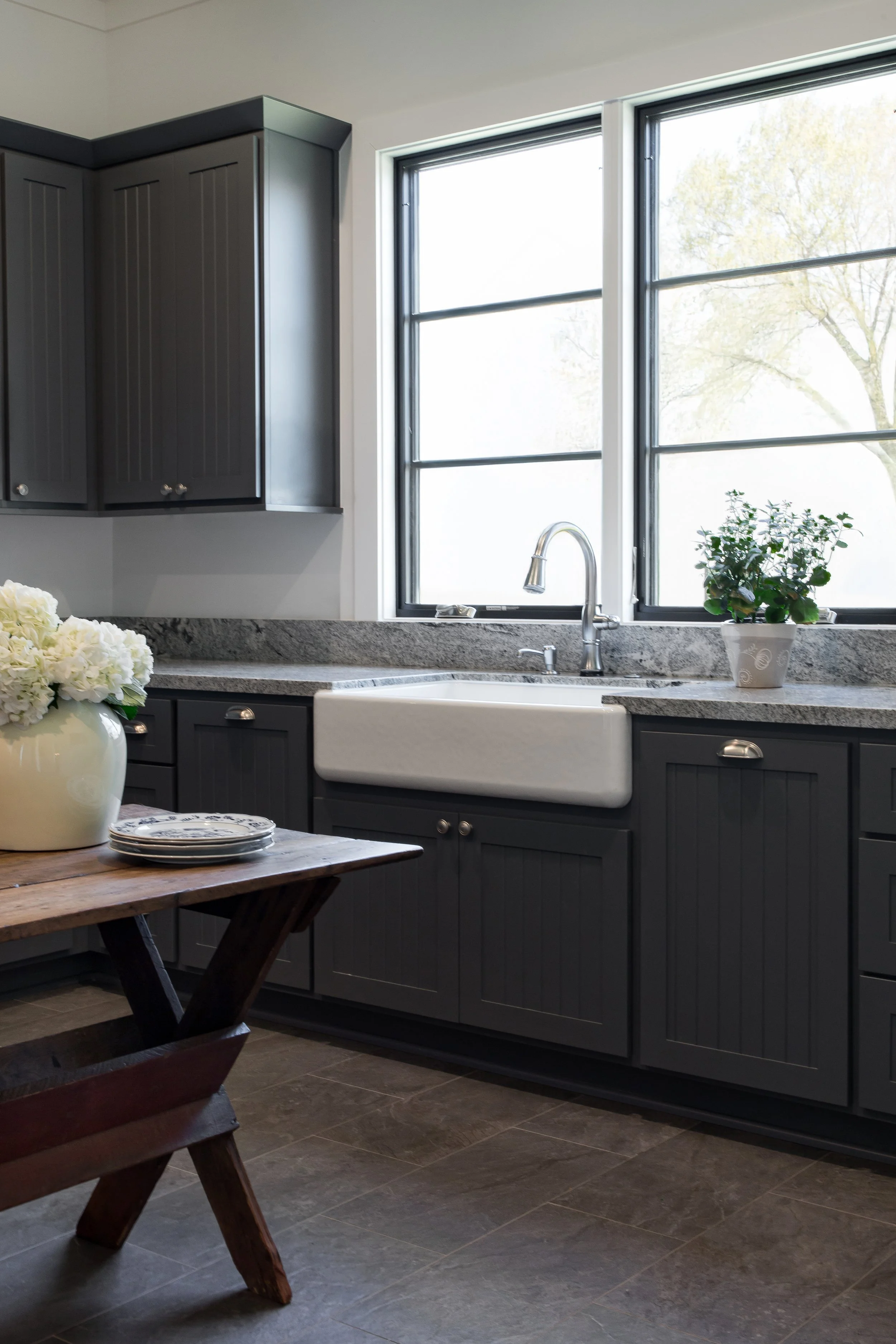 A modern kitchen with dark gray cabinets, a white farmhouse sink, granite countertop, windows overlooking greenery, decorated with a small potted plant and a vase with white flowers.