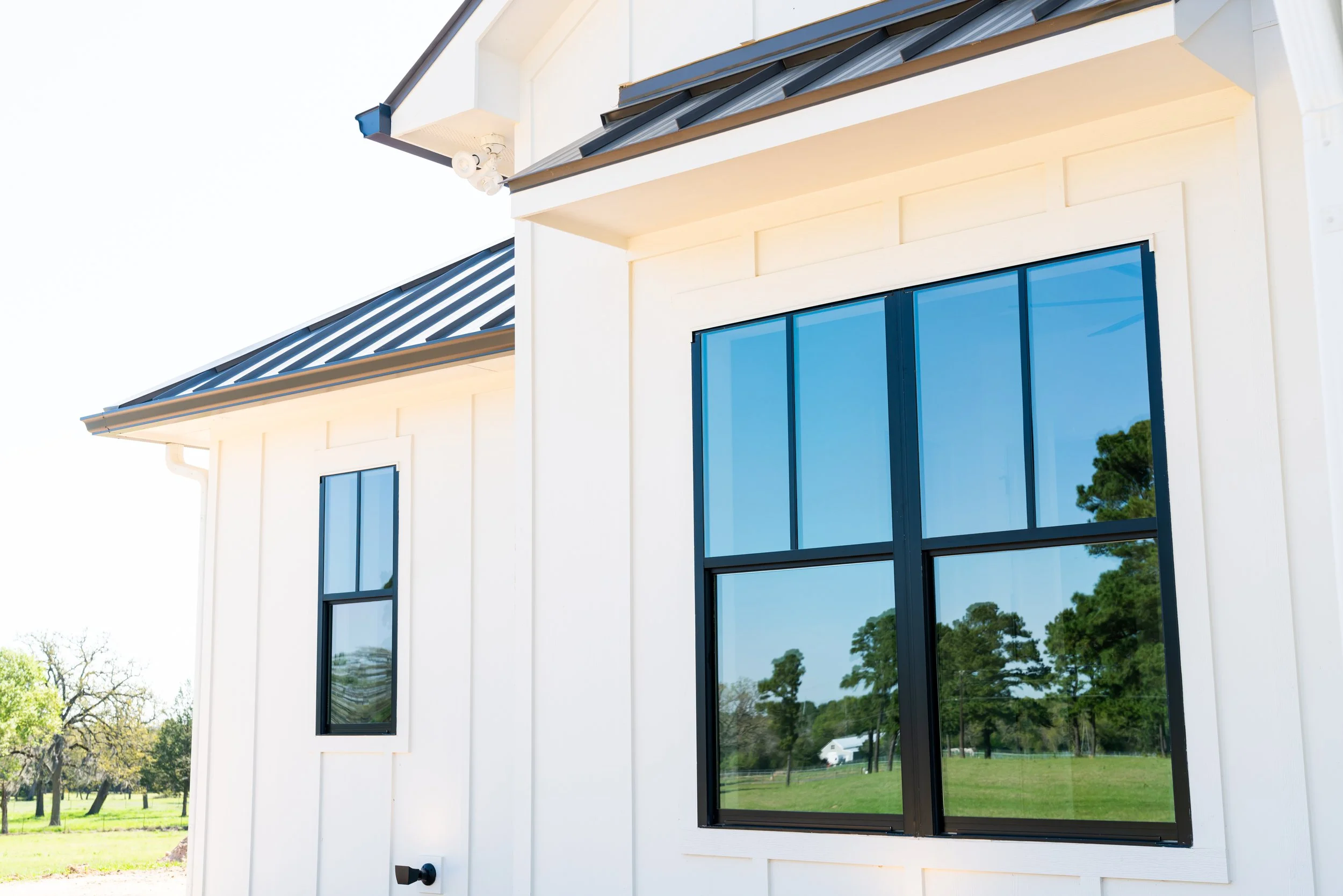 Close-up of a modern house exterior with white siding, large black-framed windows, and a metal roof, with trees and a grassy landscape reflected in the windows.