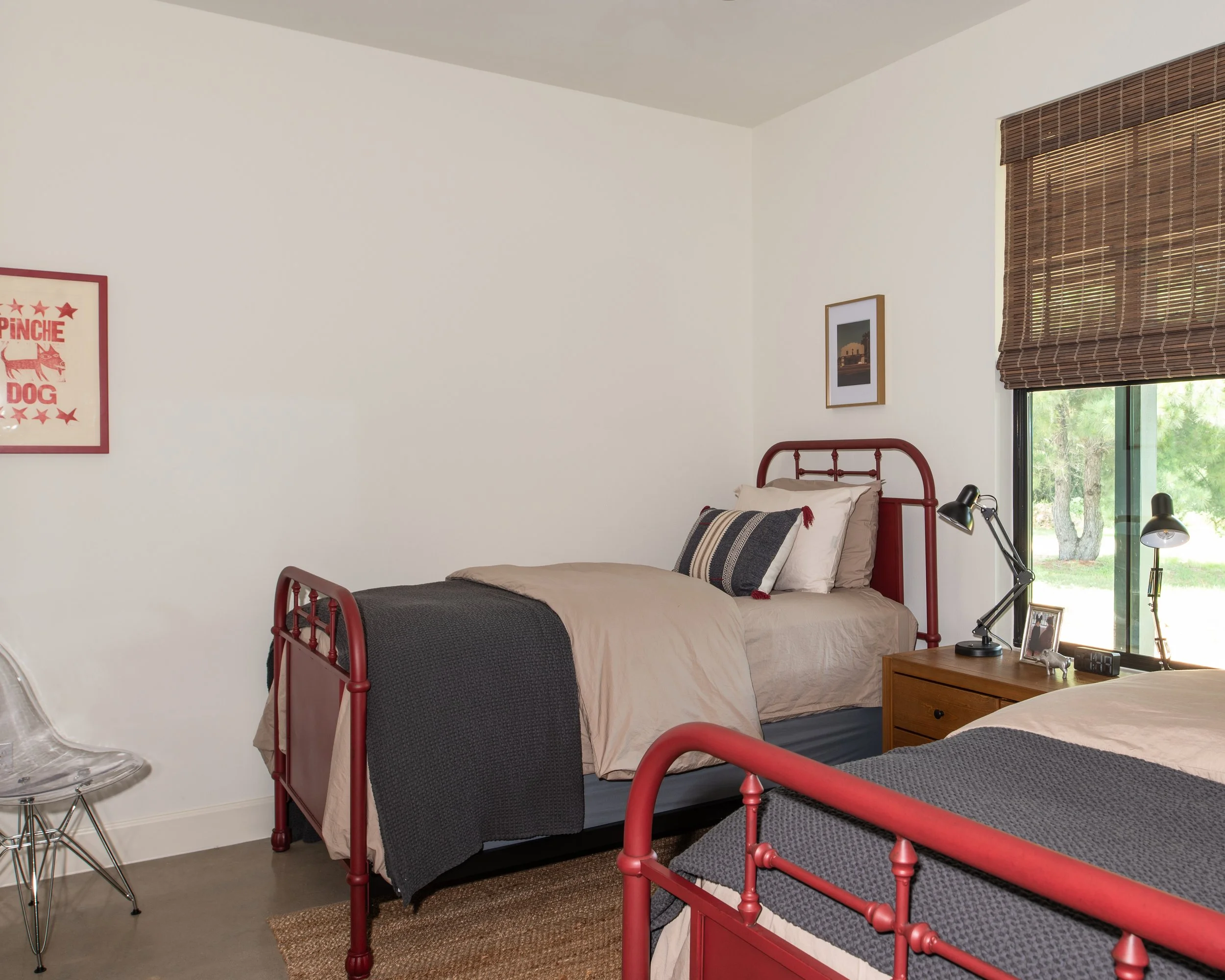A bedroom with two beds, one with a beige bedspread and black throw, the other with a dark gray bedspread. A wooden nightstand with two black desk lamps, a framed photo, and a small decorative item sits between them. There is a window with brown blin
