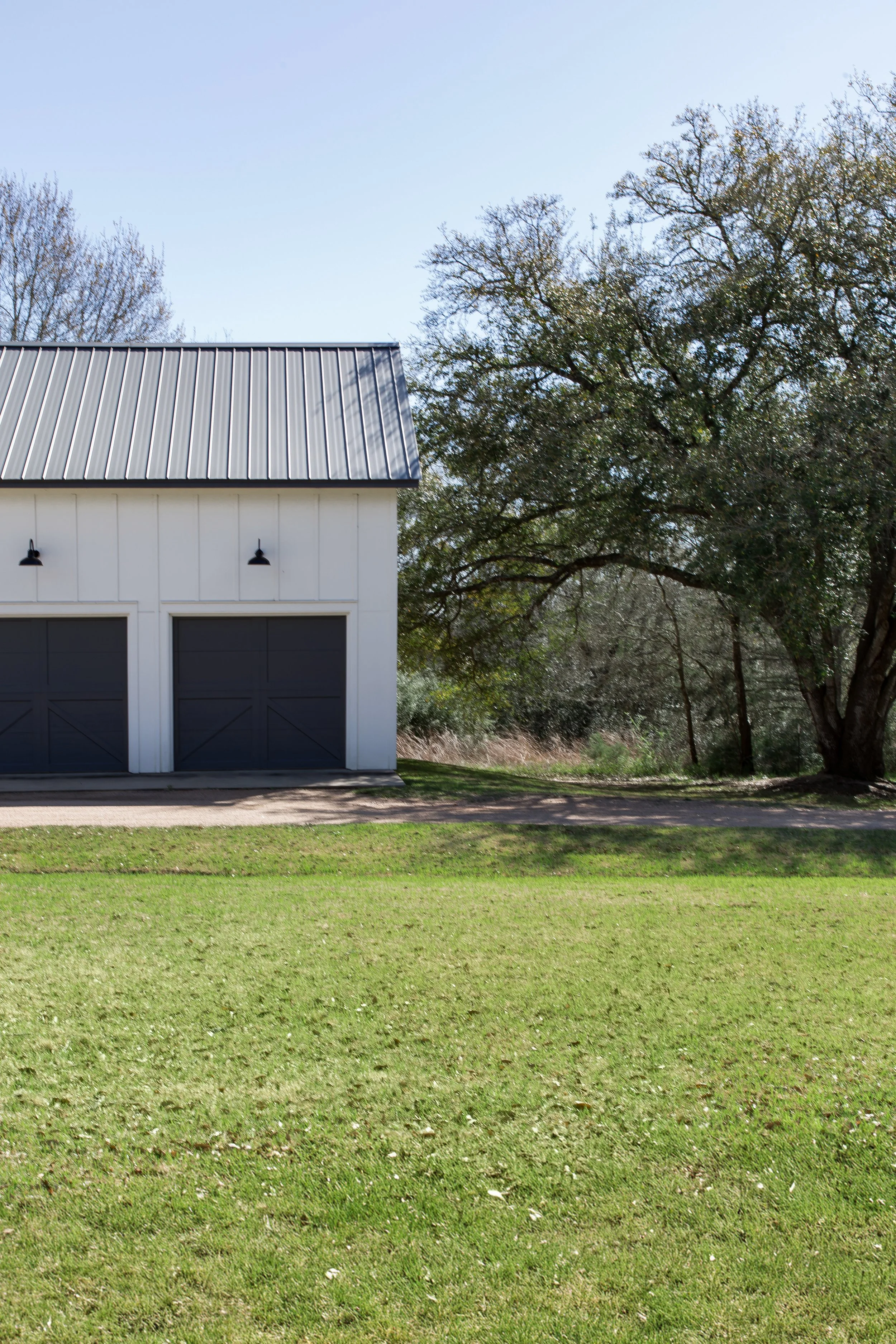 A white garage with two black doors and black outdoor lights, next to a large leafy tree on a sunny day.