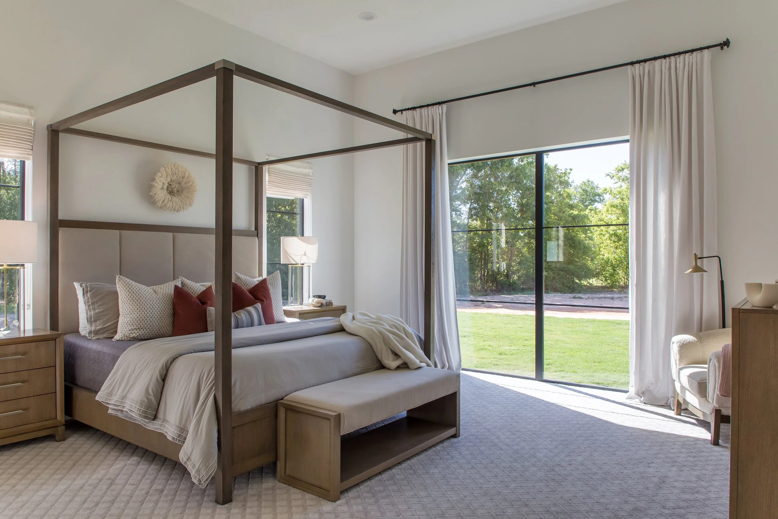 A bright bedroom with a large four-poster bed, beige upholstered headboard, and various pillows. Large sliding glass door with white curtains opens to a grassy outdoor area with trees, allowing natural sunlight into the room.
