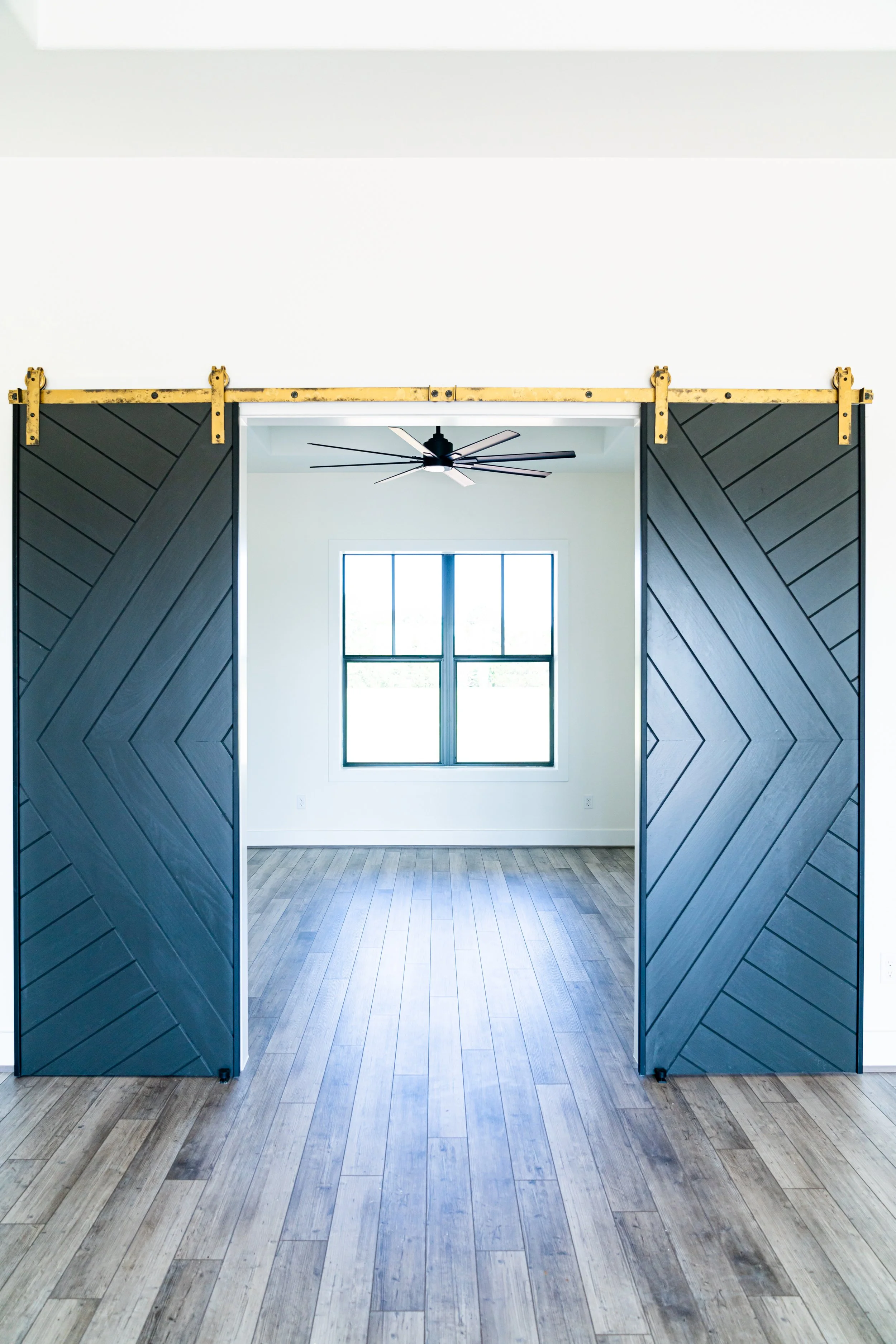 A room with black sliding barn doors, a ceiling fan, and a window with four panes, with light-colored wooden flooring and white walls.