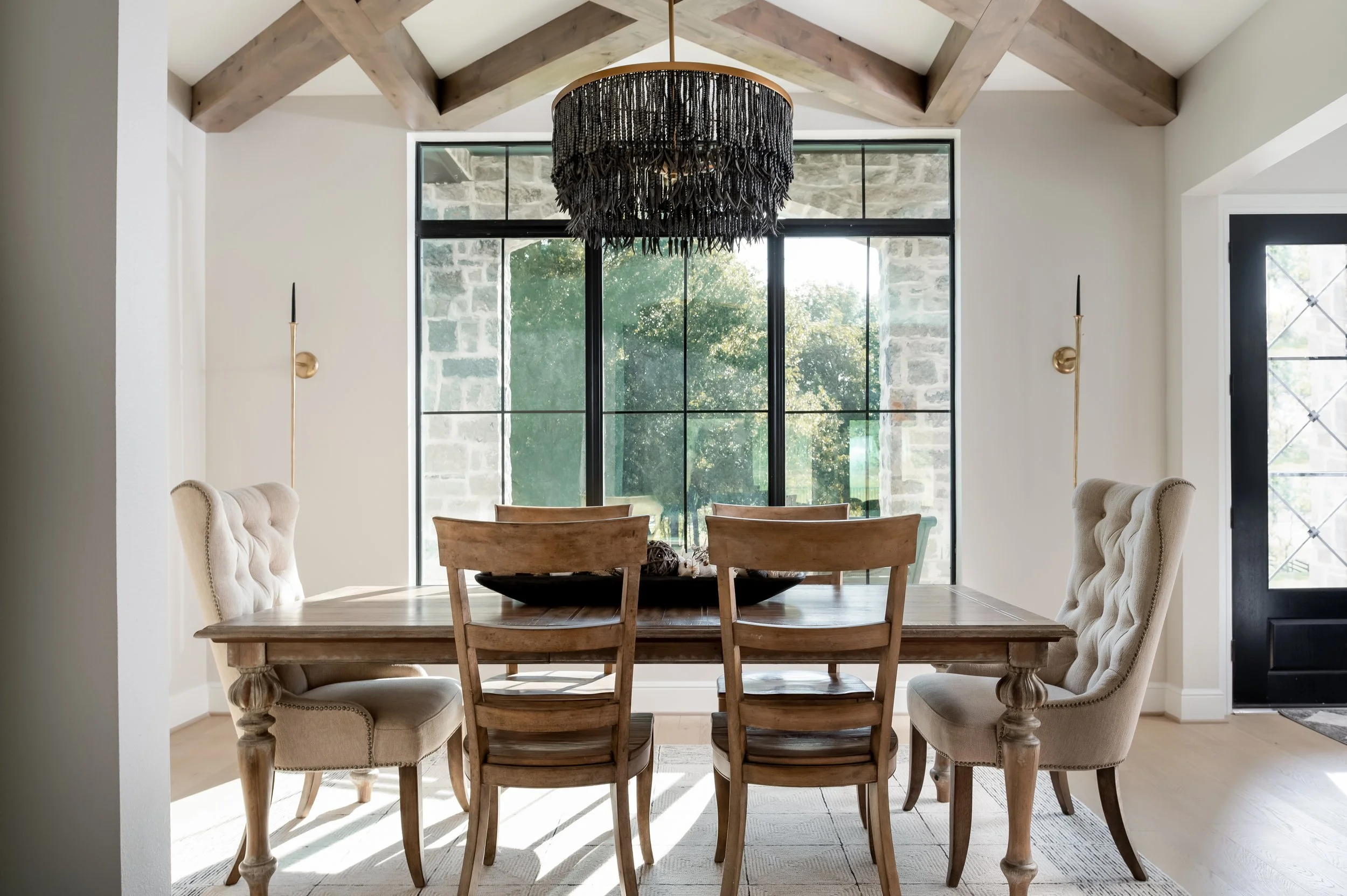 A dining room with a wooden table, six chairs, and a black chandelier hanging from a vaulted ceiling with exposed wooden beams. Large windows with black frames reveal an outdoor view of trees and stone walls.