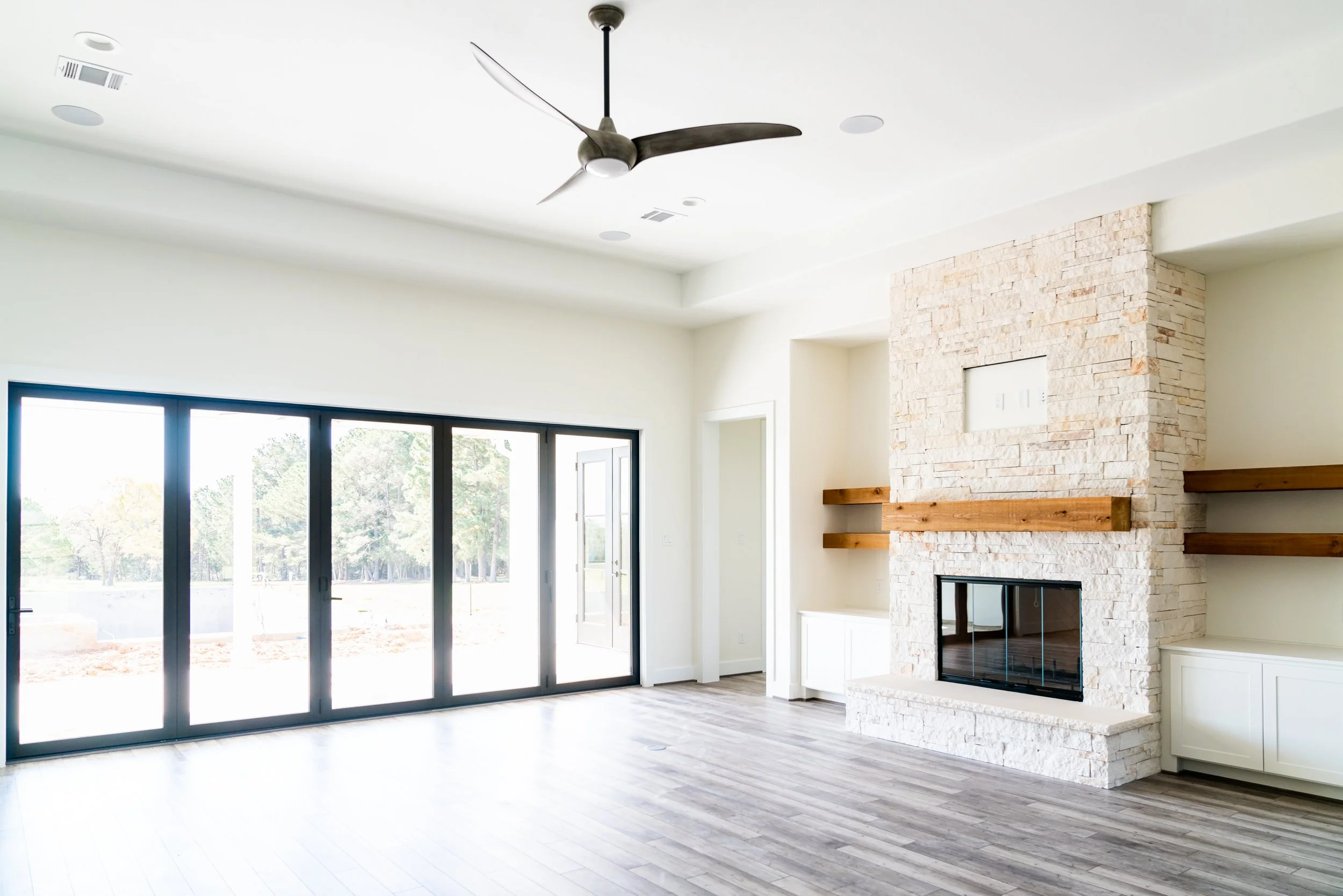 Empty living room with a modern ceiling fan, large glass sliding doors, a white stone fireplace with wooden shelves, and a view of outdoor trees.