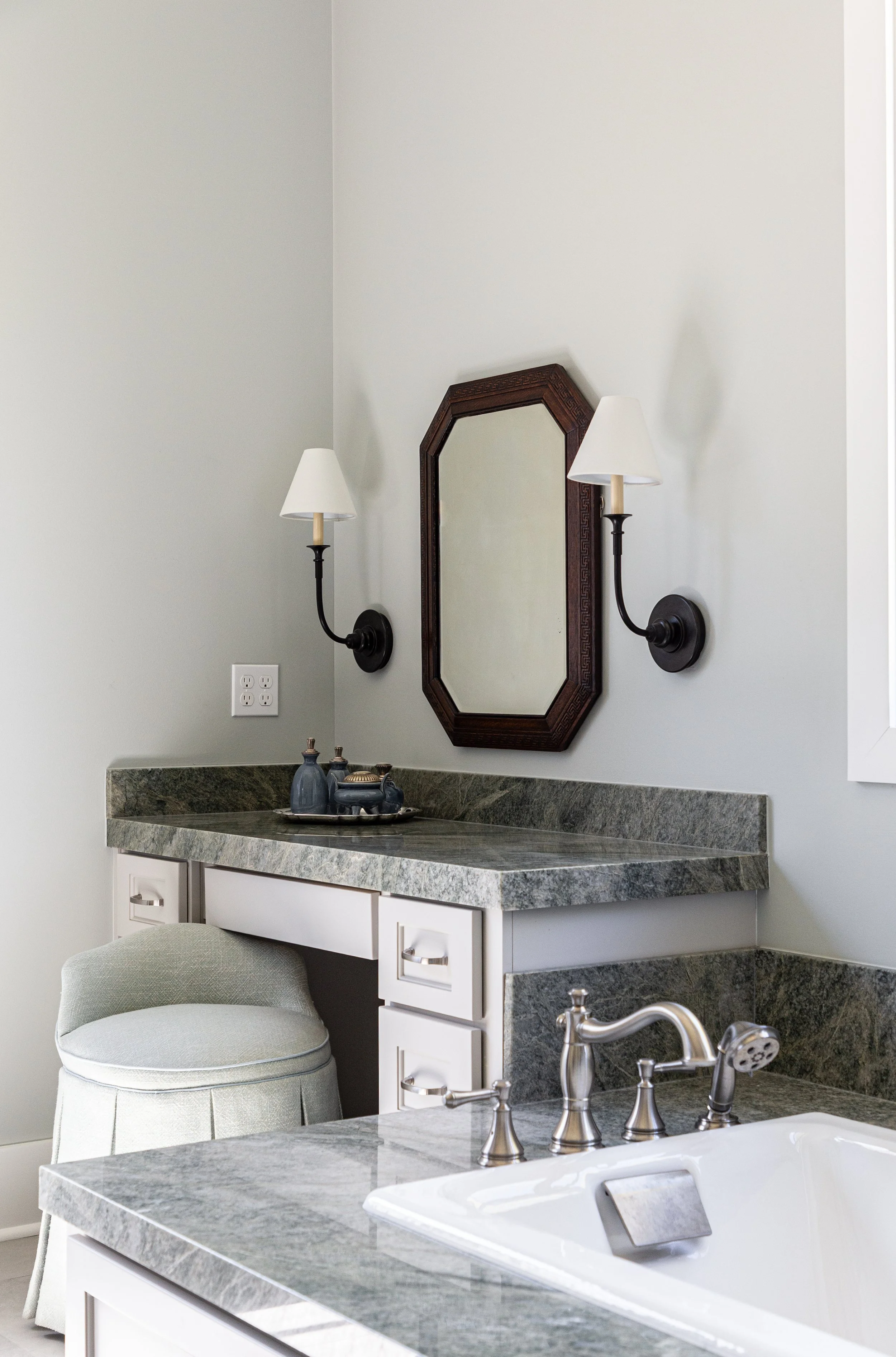 Bathroom with a granite vanity, a mirror with a dark wooden frame, two wall-mounted lamps, a small stack of decorative bottles, a cushioned stool, and a bathtub with a chrome faucet.