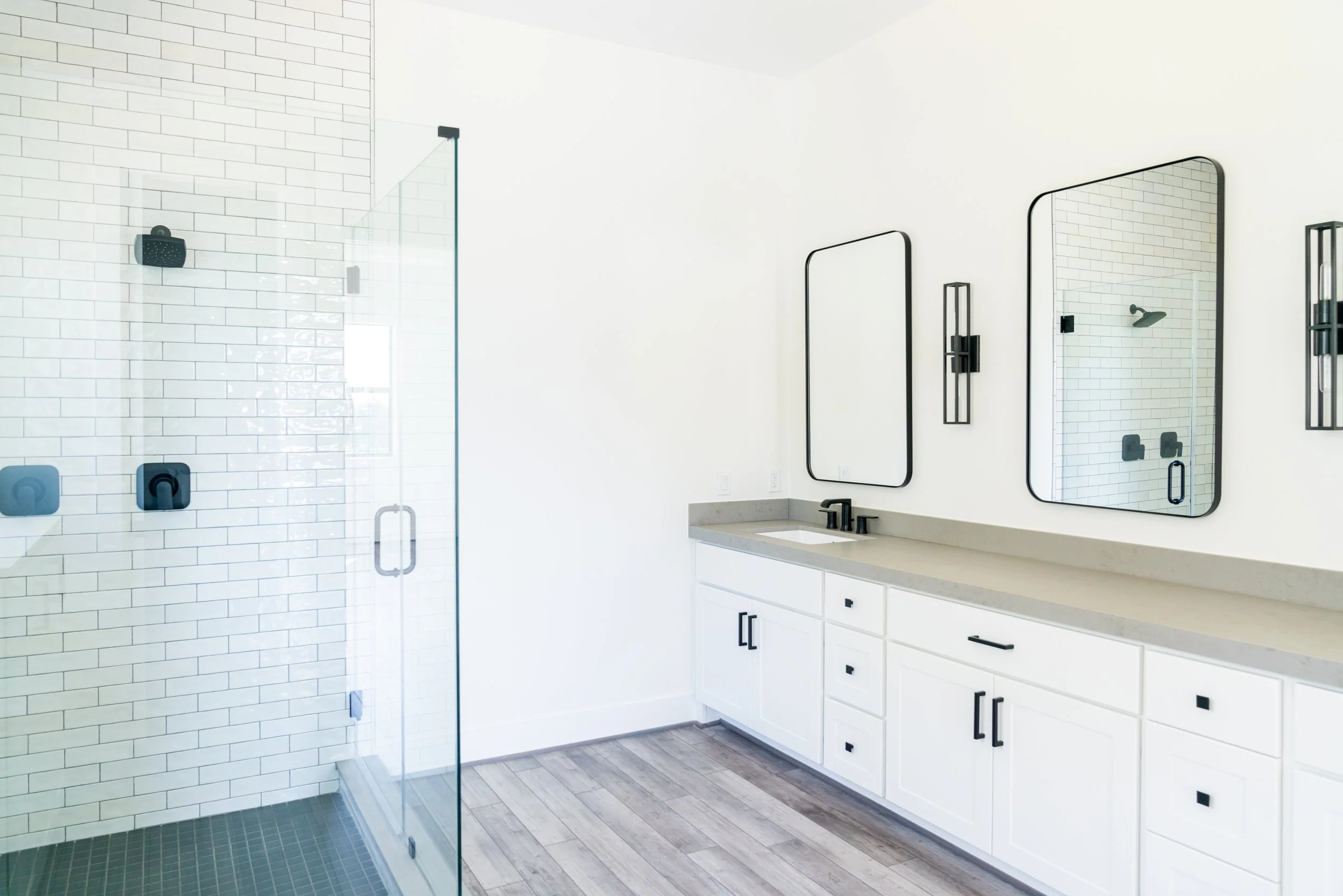 Modern bathroom featuring a walk-in shower with white subway tile walls, black fixtures, and a glass door, alongside a white vanity with two mirrors and black accents.