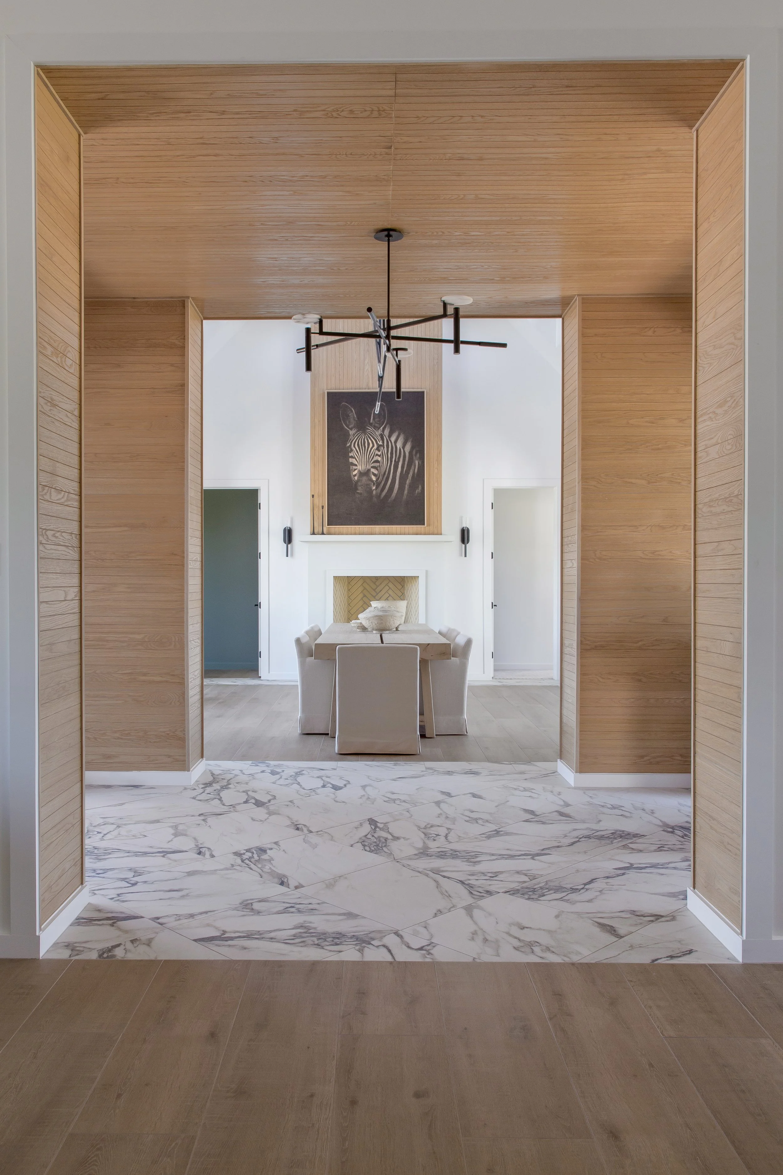 View of a dining room with a modern chandelier, a large zebra painting, a fireplace, a wooden dining table with chairs, and a marble and wooden floor.