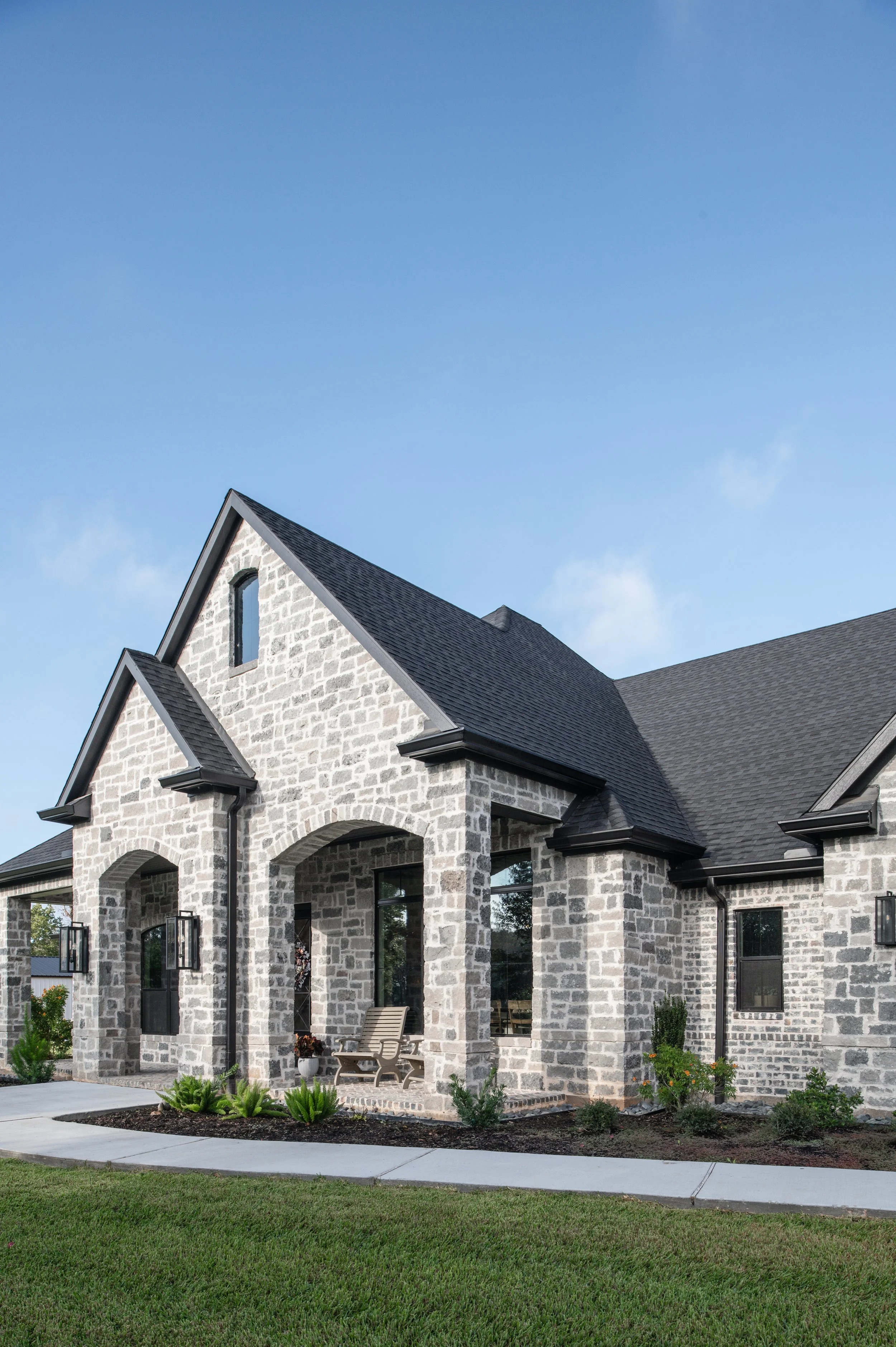 A modern house with a stone exterior, black roof, arched entryway, and large windows, surrounded by a green lawn and sidewalk.