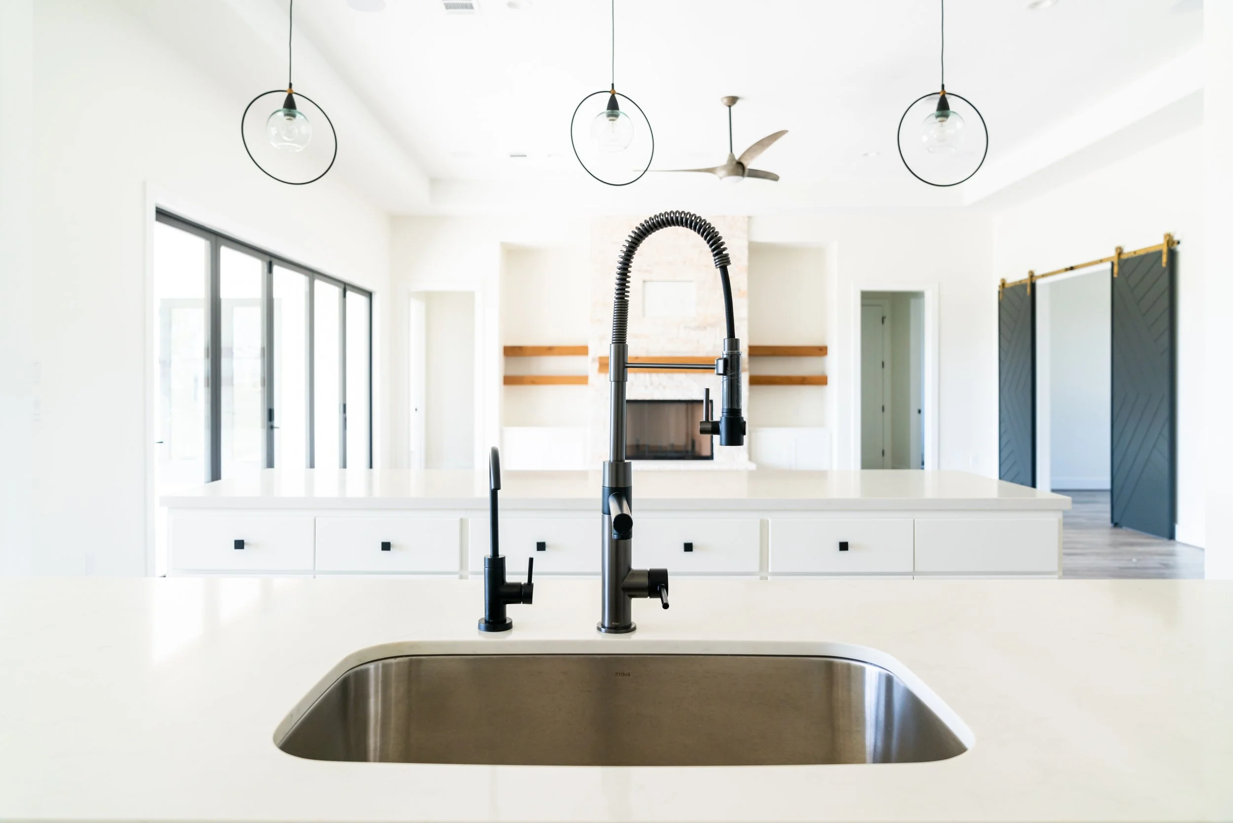 Modern kitchen with white island, stainless steel sink, black faucet, pendant lights, sliding glass door, fireplace, and sliding barn doors.