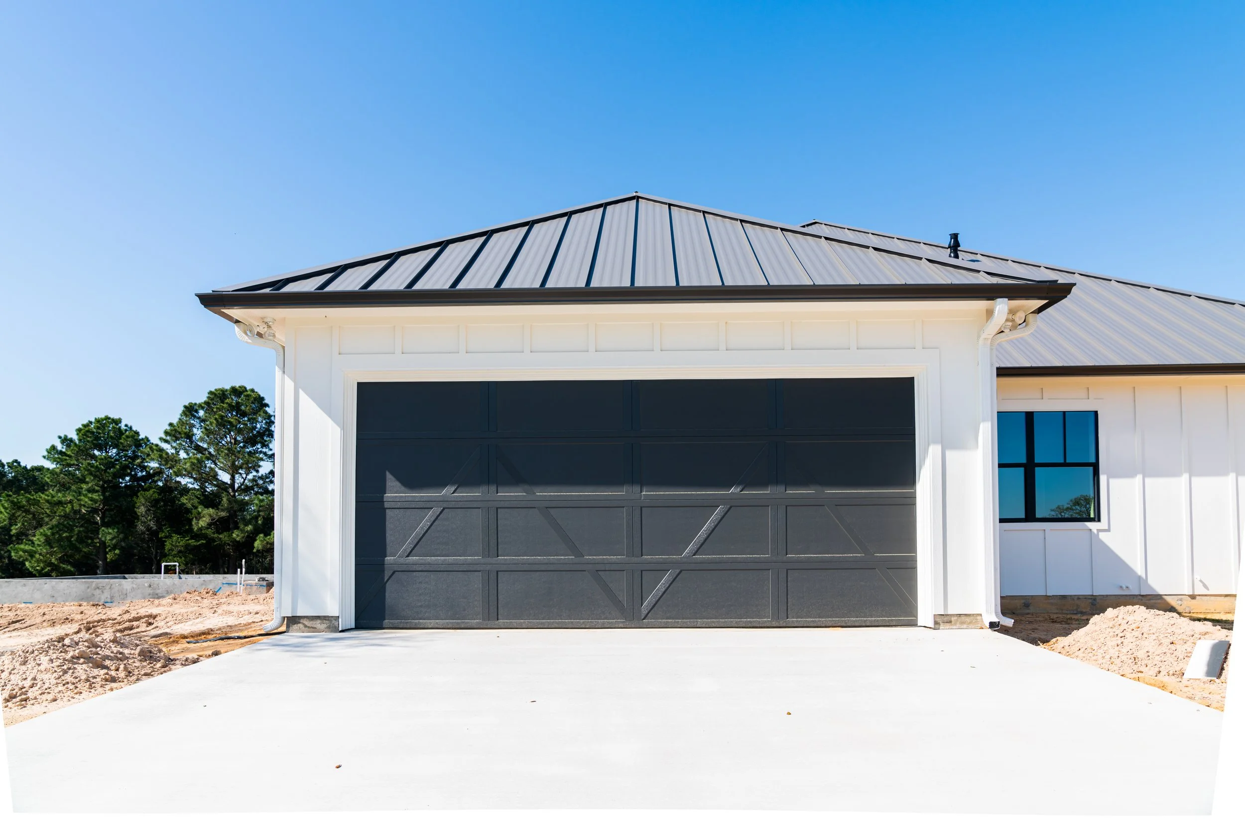 New house with black garage door, white exterior, metal roof, and a window with reflection of trees, under a clear blue sky.