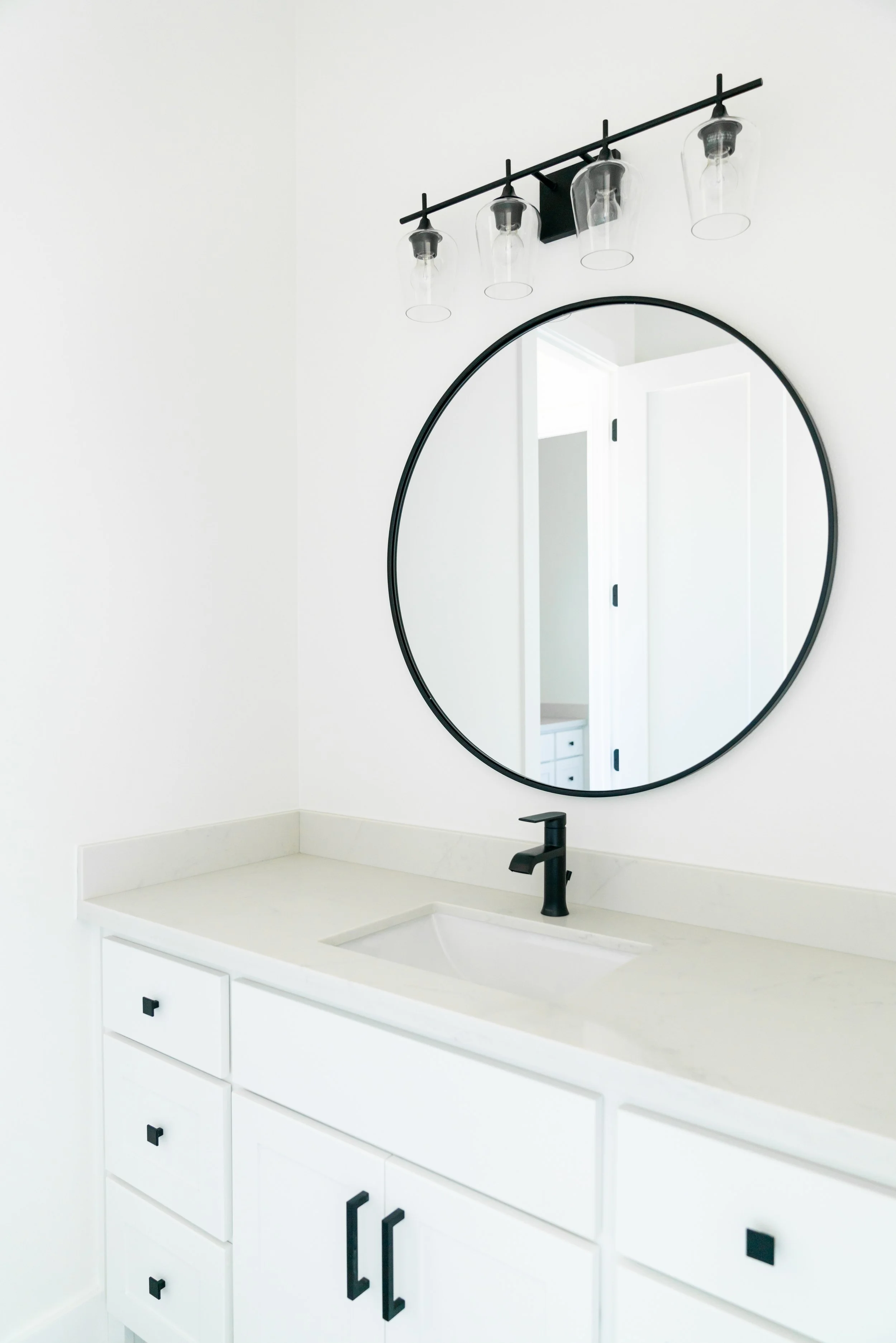 White bathroom vanity with black fixtures, a large round mirror, and a black light fixture with four clear glass shades.