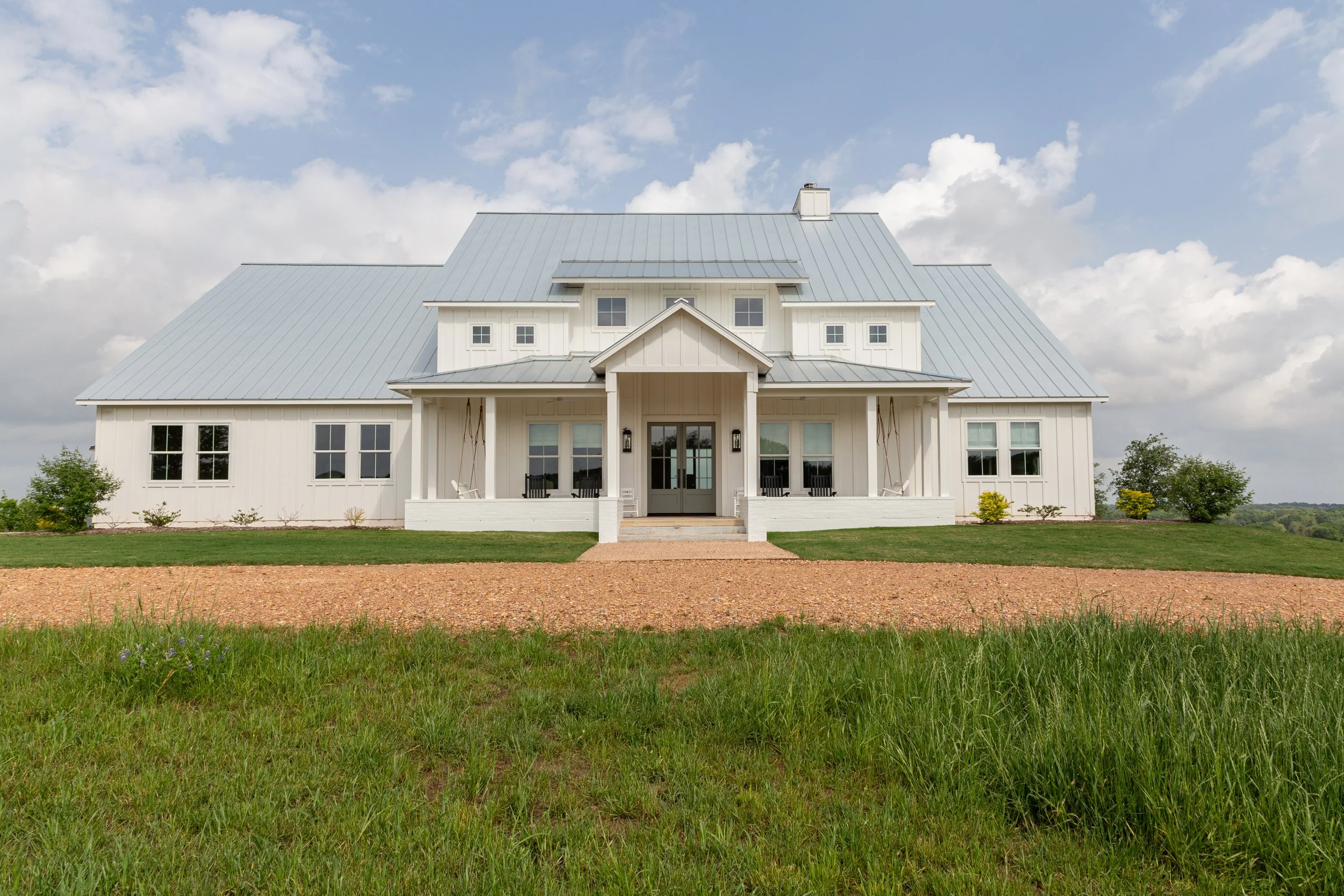 A white farmhouse-style house with metal roof, front porch with hanging swings, stairs leading up to doors, surrounded by green grass and trees, under a partly cloudy sky.