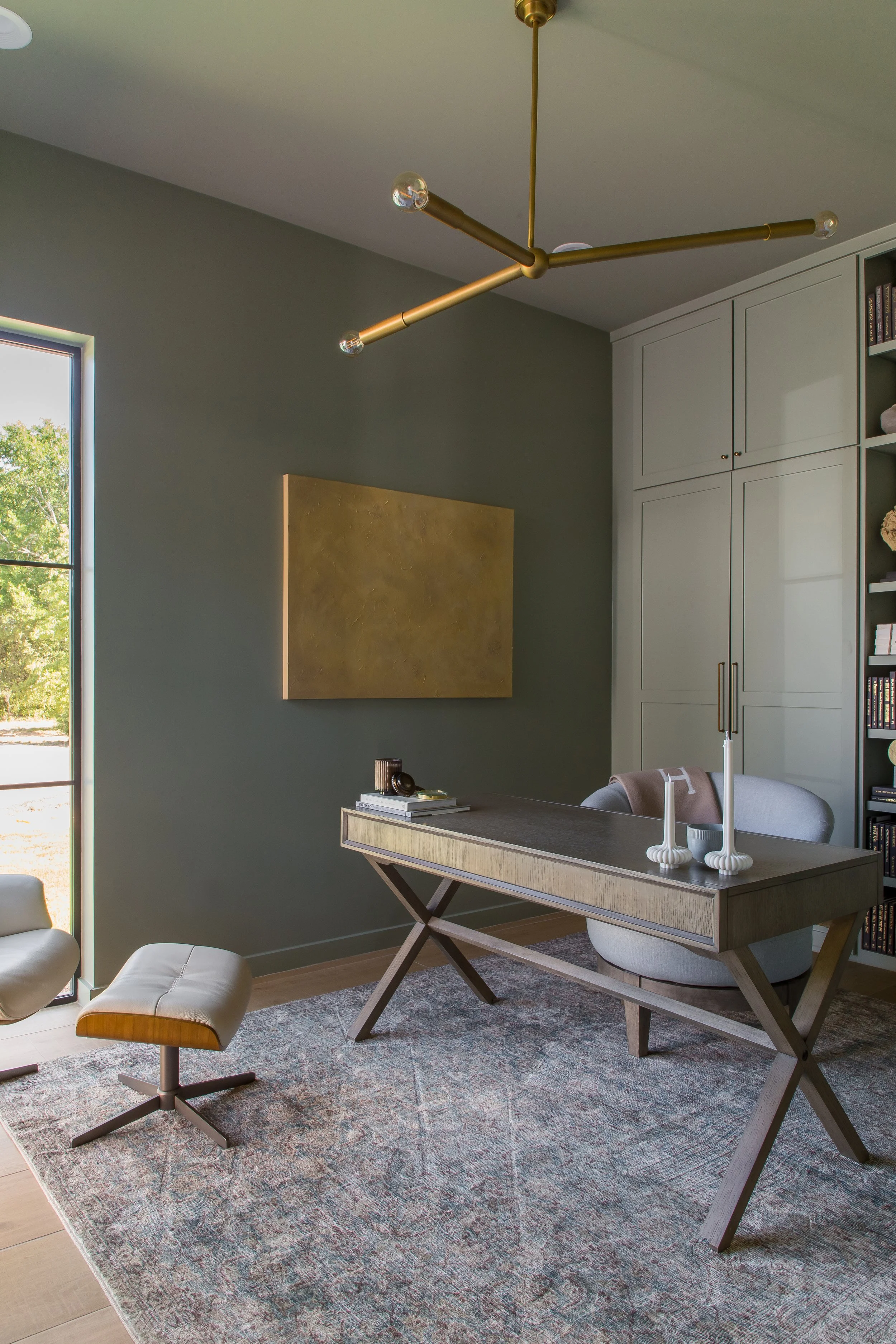 Modern home office with gray walls, a large window, a wooden desk with angled legs, a light gray swivel chair, and a patterned area rug. A gold abstract wall art piece hangs on the wall, and a gold multi-arm ceiling light fixture is overhead.