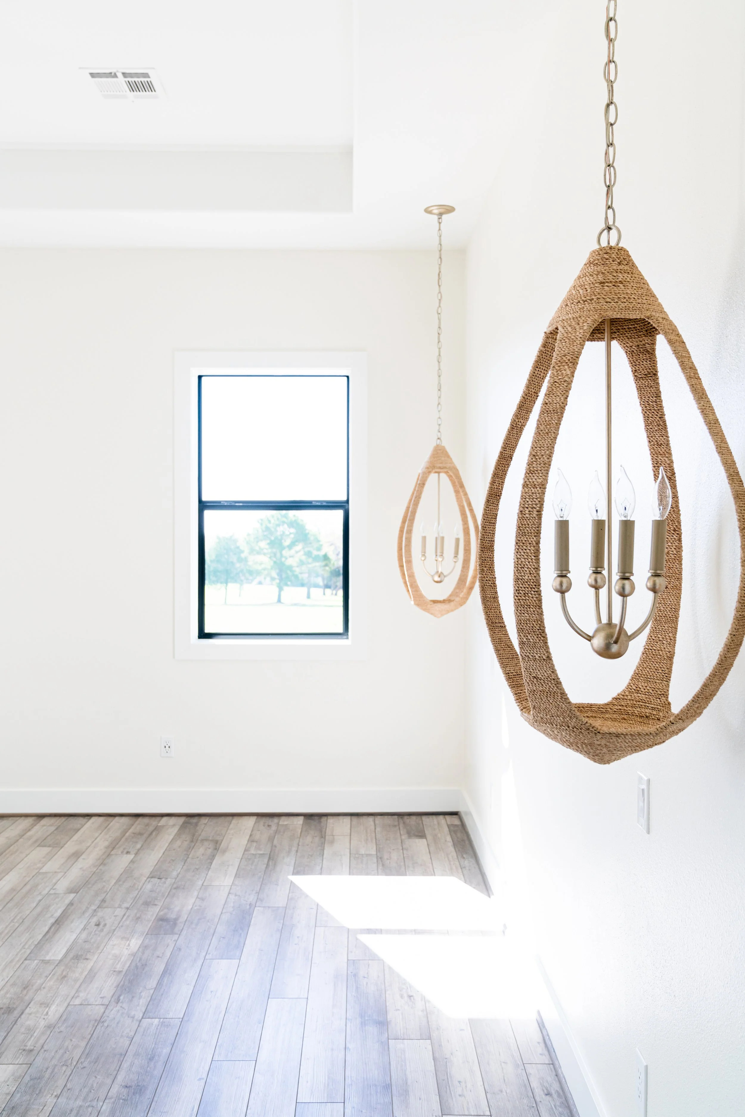 Two modern chandelier light fixtures with woven rope surrounds hanging in an empty room with white walls, a window, and wood flooring.