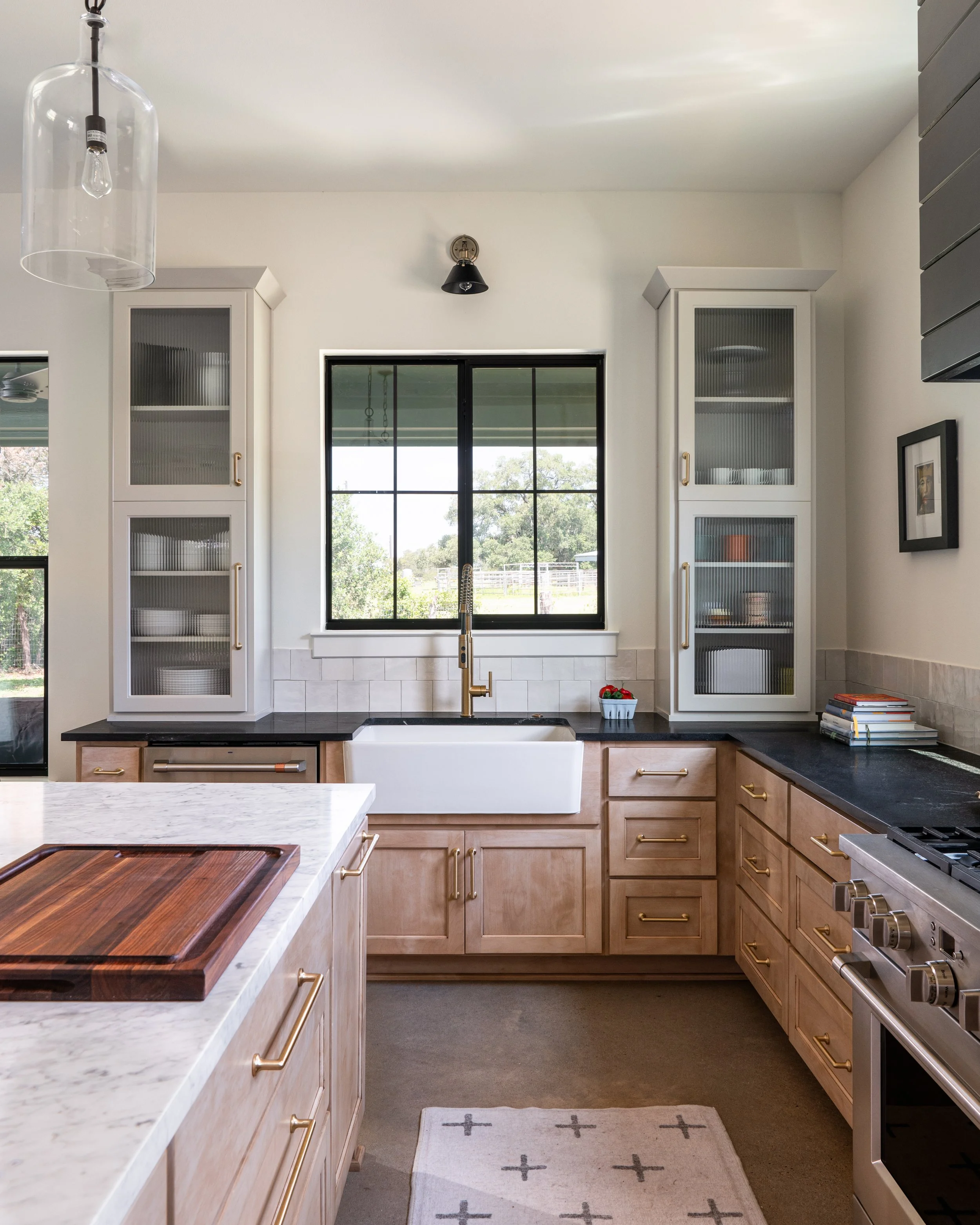 Kitchen with white cabinets, black countertops, a window above the farmhouse sink, a cutting board on the island, and a gray and white rug on the floor.