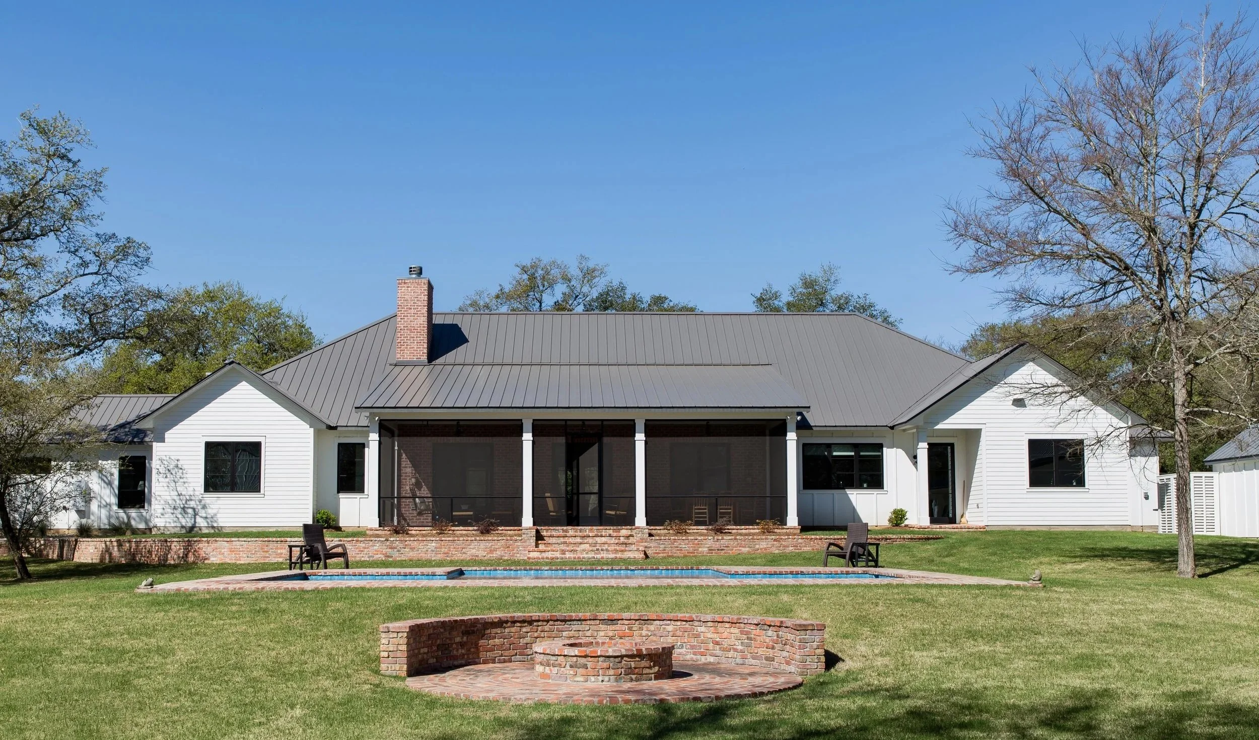 A large white house with a gray metal roof and a brick chimney, surrounded by trees and a grassy yard. There is a brick fire pit and a small swimming pool in the yard, with a couple of chairs near the pool.