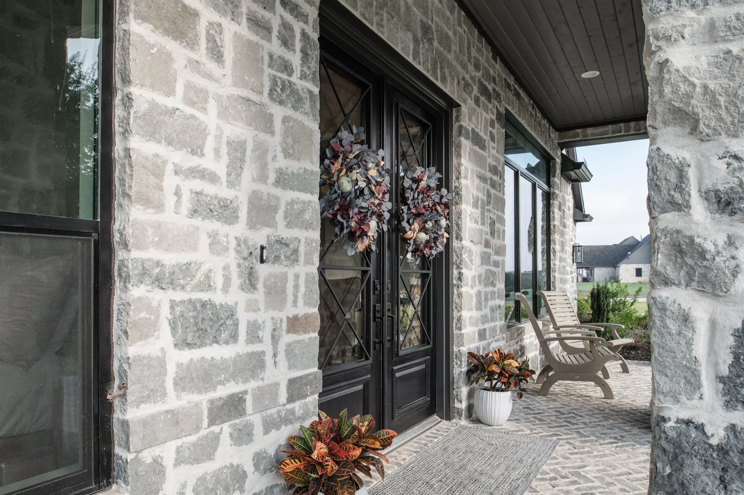 Front porch of a house with stone walls, black security doors decorated with fall-themed wreaths, and potted plants near the entrance. There are two wooden rocking chairs on the brick and stone porch, with a view of neighboring houses and green lawns