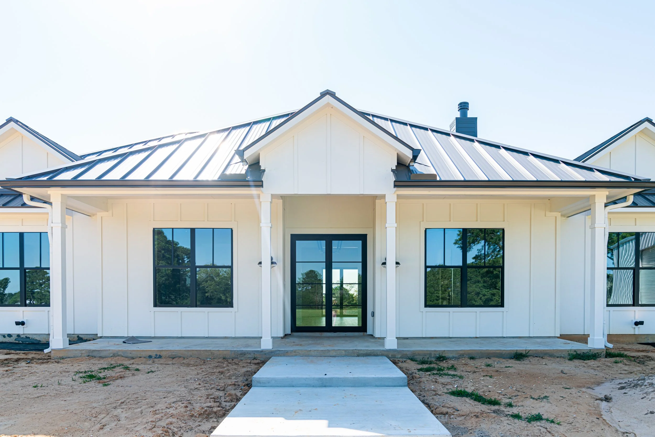 New white house with black roof, large windows, and a concrete walkway leading to the front door, under a clear blue sky.