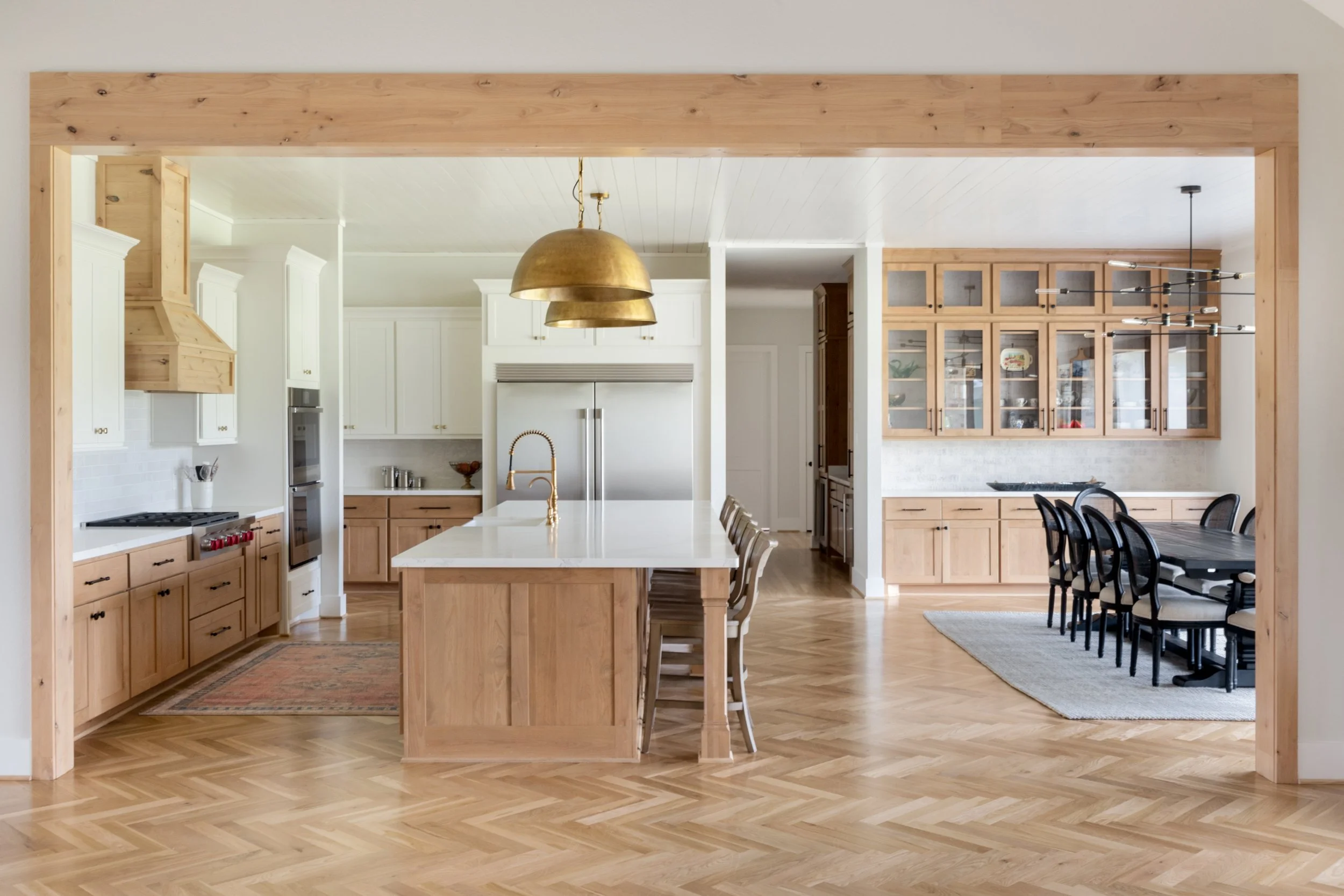 Open-concept kitchen and dining area with natural wood cabinetry, white walls, and wooden flooring, featuring a kitchen island with a gold faucet and pendant lights, and a dining table with chairs.