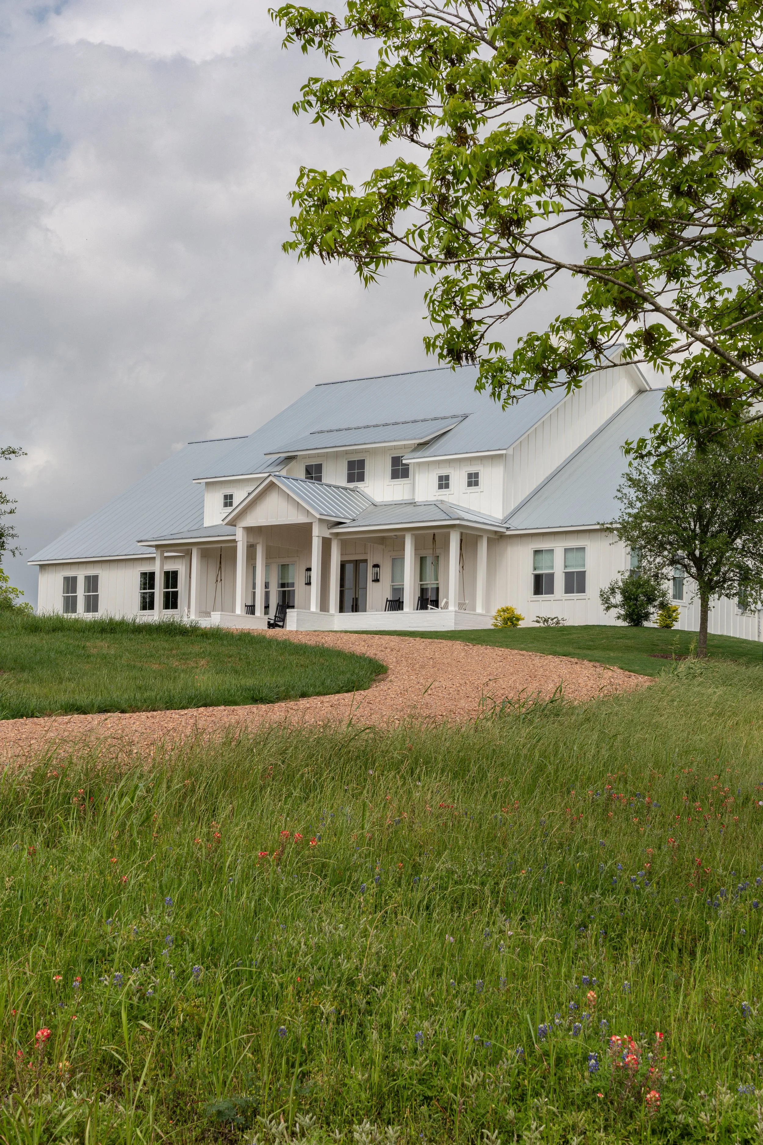 A white farmhouse-style house with a metal roof, surrounded by green grass and trees, under a cloudy sky.