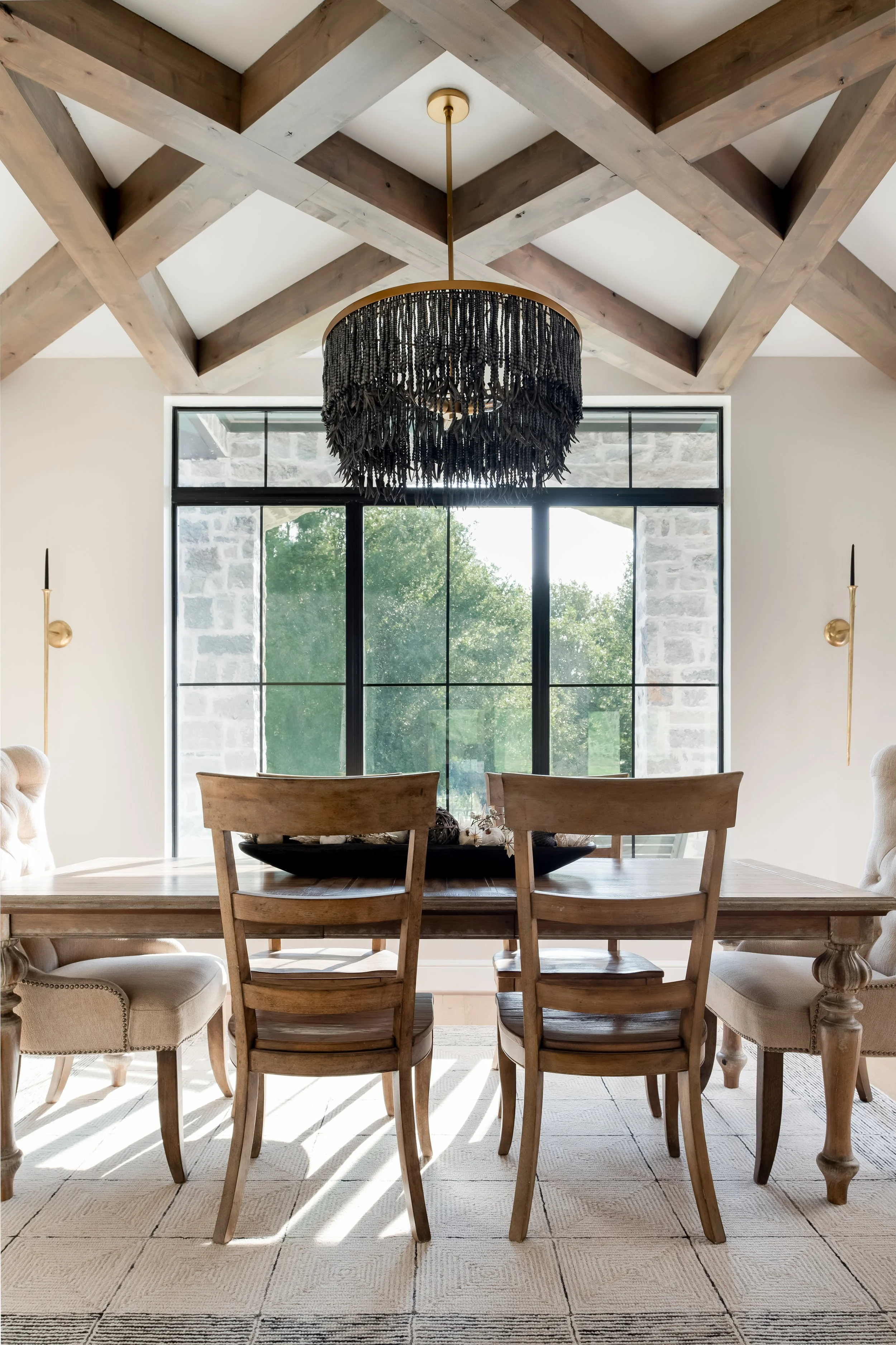 A dining room with a wooden table, six chairs, and a large black decorative chandelier hanging from a beamed ceiling. A large window with a view of trees outside is in the background, with sunlight creating shadows on the tiled floor.