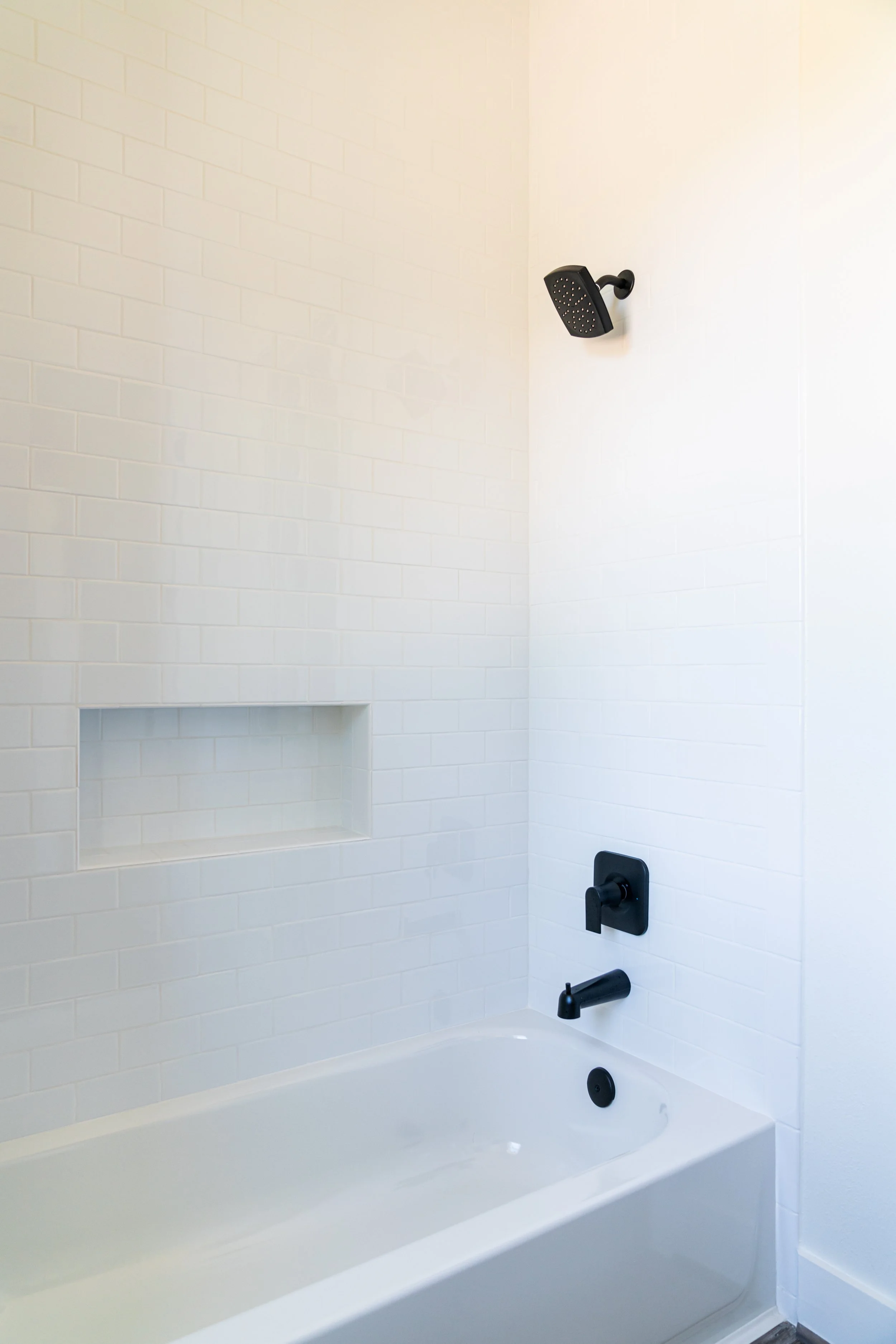 Black showerhead, faucet, and control handle on white tiled bathroom wall above white bathtub.