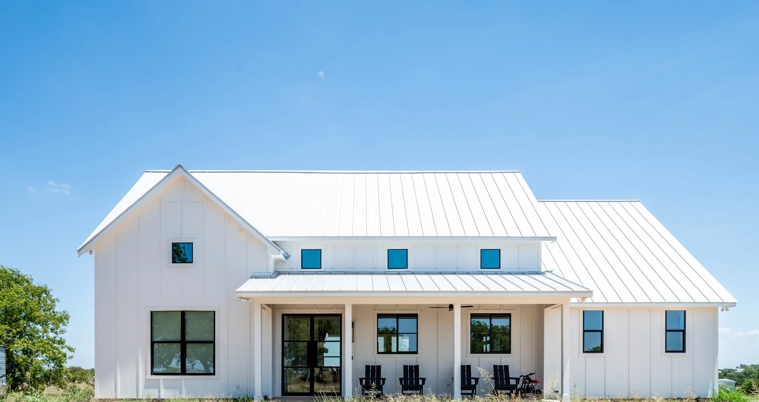 A modern white house with a metal roof, multiple windows, a porch with black chairs and a small red motorcycle, surrounded by a grassy yard and trees under a clear blue sky.