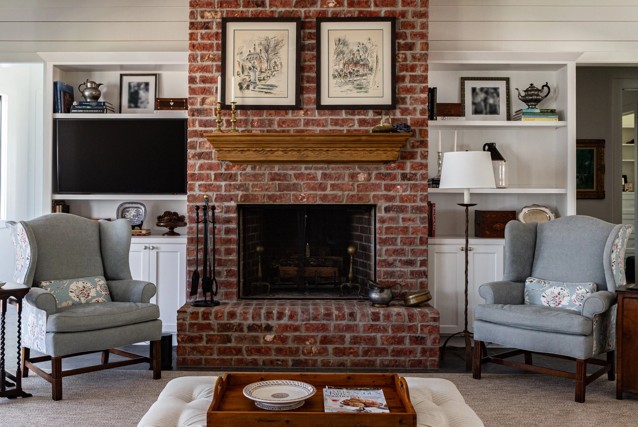 Living room with a brick fireplace, framed artwork above the mantel, two upholstered armchairs with patterned pillows, built-in white shelves with books and decorative items, a flat-screen TV, a standing lamp, and a coffee table with a magazine and d