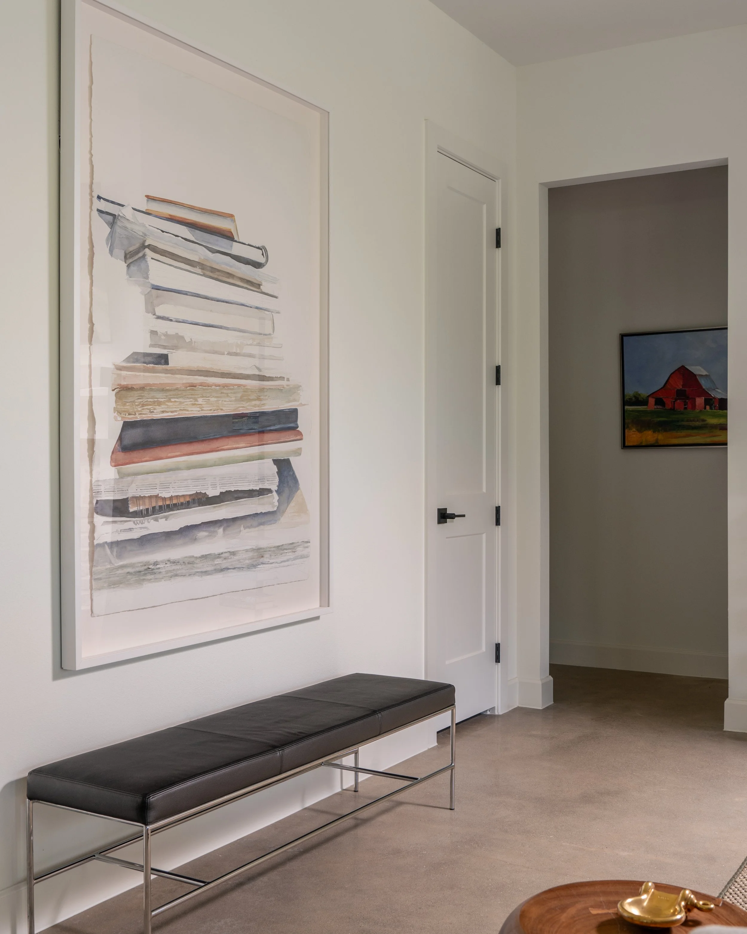 Interior of a modern home with a white wall, a large framed watercolor painting of stacked books, a black bench with metal legs, and a doorway leading to another room with a landscape painting of a barn.