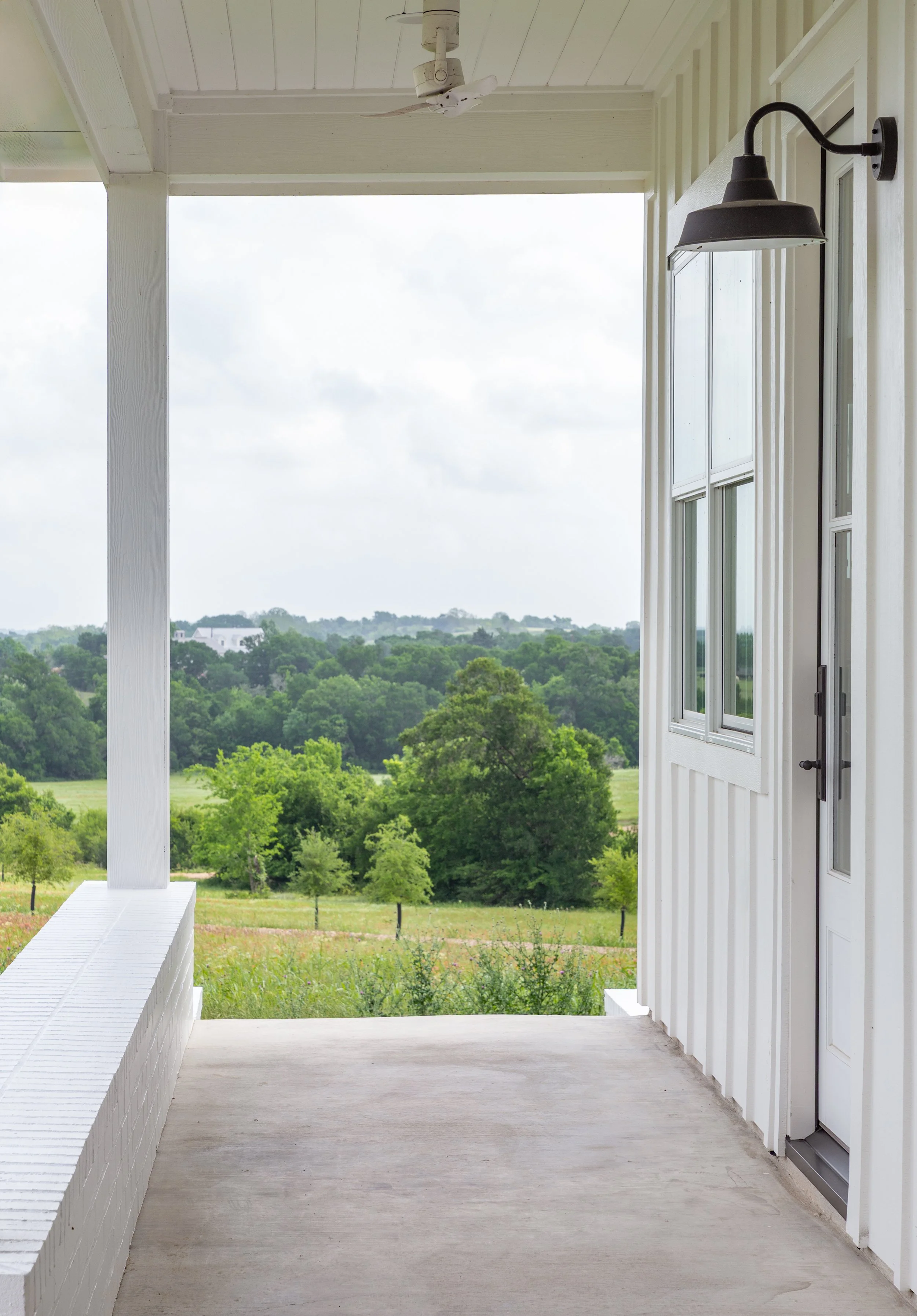 A porch with a white railing and wall, black outdoor wall light, and a ceiling fan, overlooking a green landscape with trees and cloudy sky.