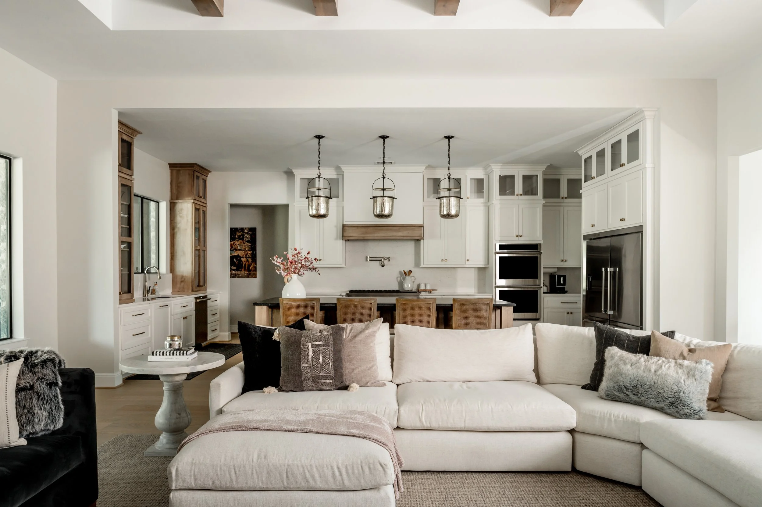 Living room with a white sectional sofa, decorative pillows, a textured blanket, a small round side table with a gray base, and a black armchair. Open kitchen in the background with white cabinets, pendant lights, a large refrigerator, and a dining a