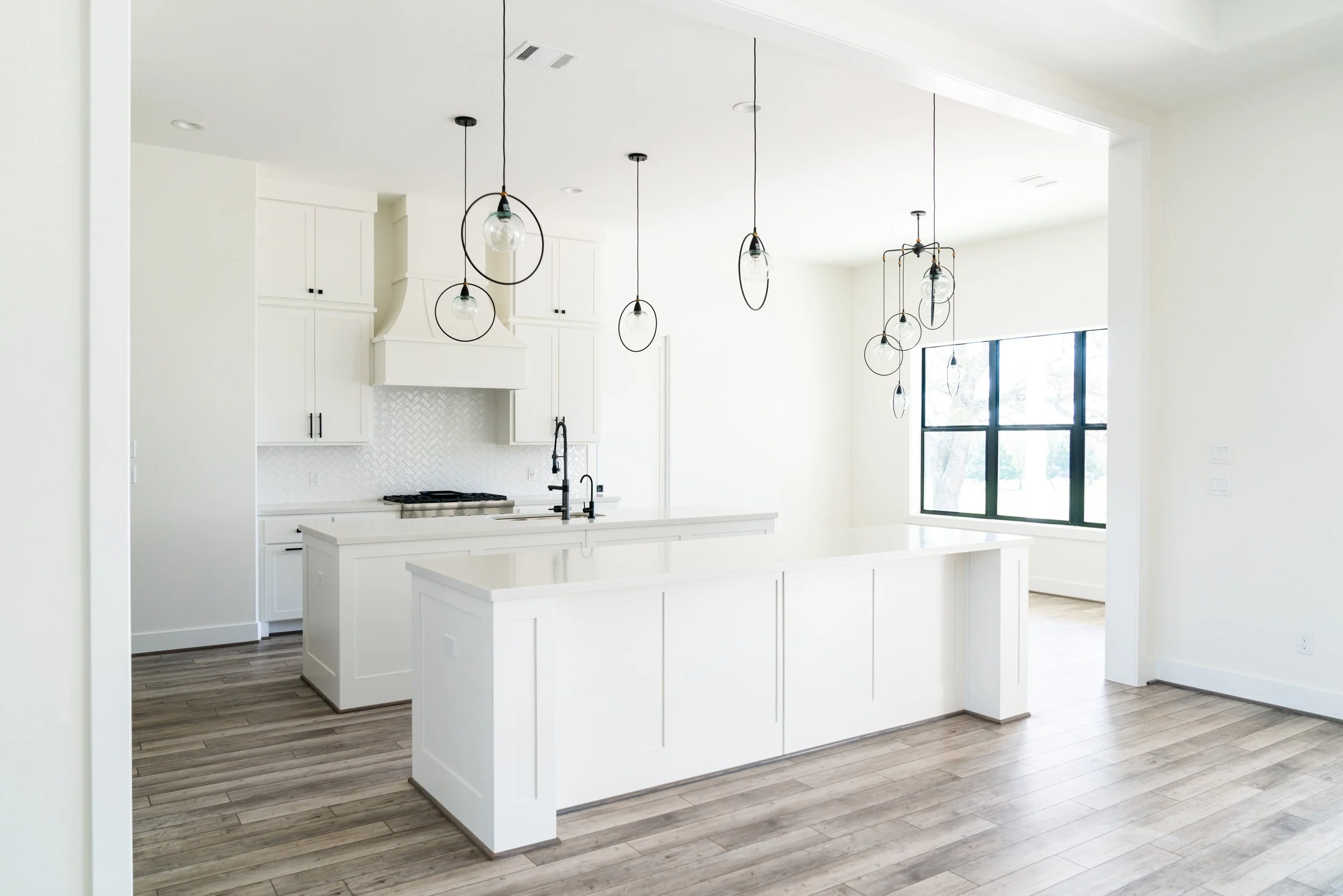 A bright, modern kitchen with white cabinets, a central island, and minimalist pendant lighting. Large window with black frames lets in natural light, and light wood flooring completes the space.