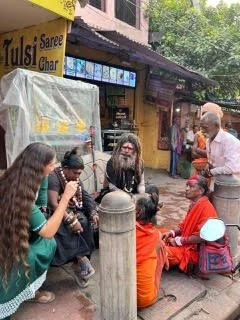 A woman with long dark hair, wearing traditional Indian attire with a saree and jewelry, smiling and holding her hands in a namaste gesture.