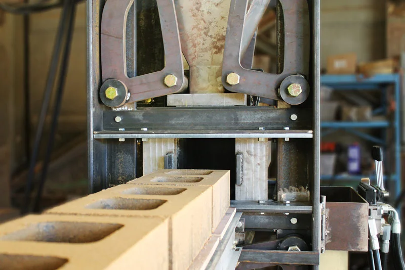 The first version of our new high compression block machine uses levers and rollers to amplify compressive force by a factor of four. The end gates close off the compression chamber during block fabrication then slide open to allow the finished block to land on the off-loading rollers. The end gate doubles as one of the four sides of the mold. The back end of the mold similarly doubles as an extractor to push the block out of the core. In this image, the end gates are not connected to their hydraulic cylinders.