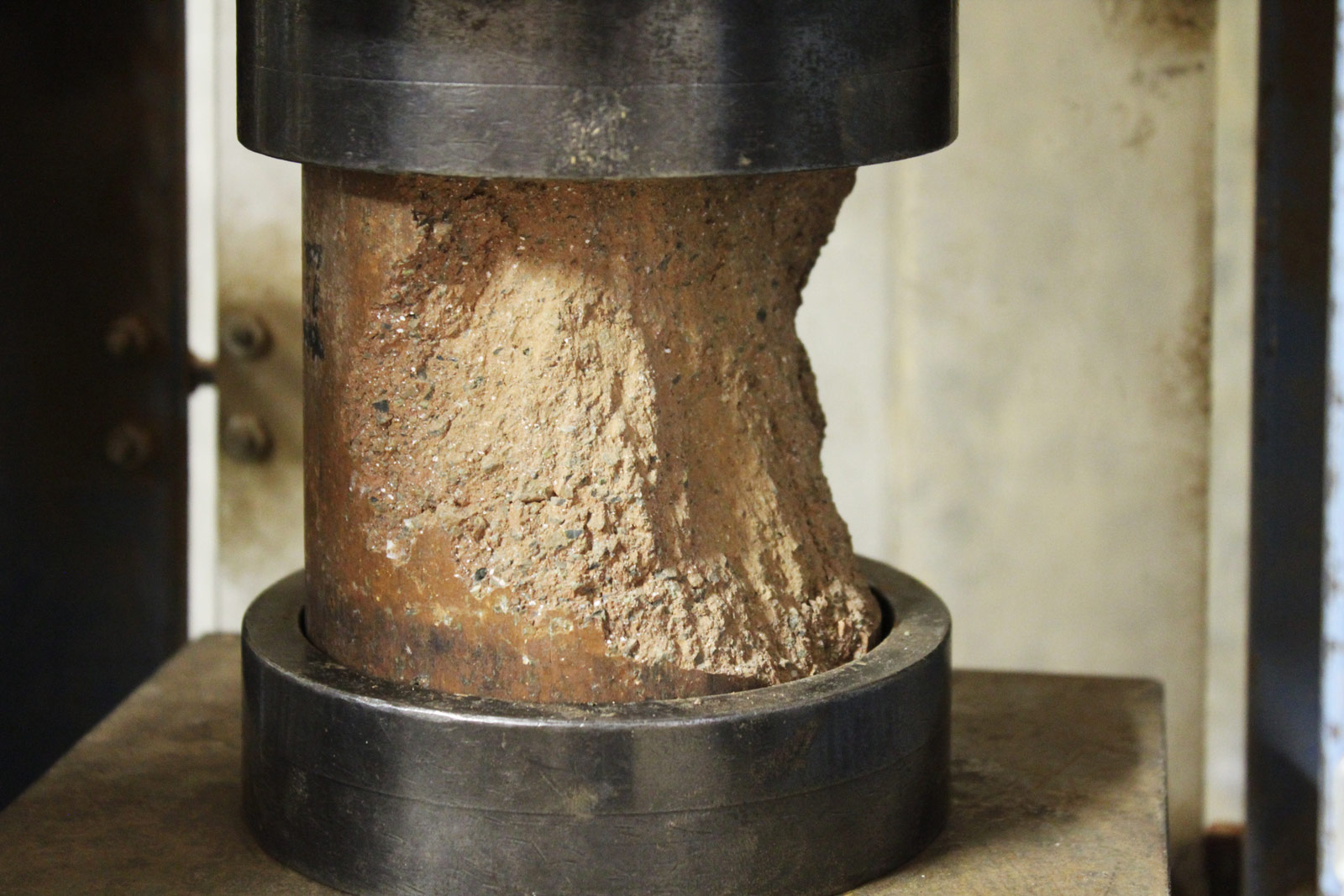 Compressive strength testing of a Watershed Materials’ geopolymer test specimen. This sample, sandwiched between two steel caps equipped with neoprene pads to distribute loads, finally failed after being loaded with 100,000 pounds of force. This load equates to a compressive strength of 7,000 pounds per square inch, 350% stronger than the requirement for typical concrete masonry.