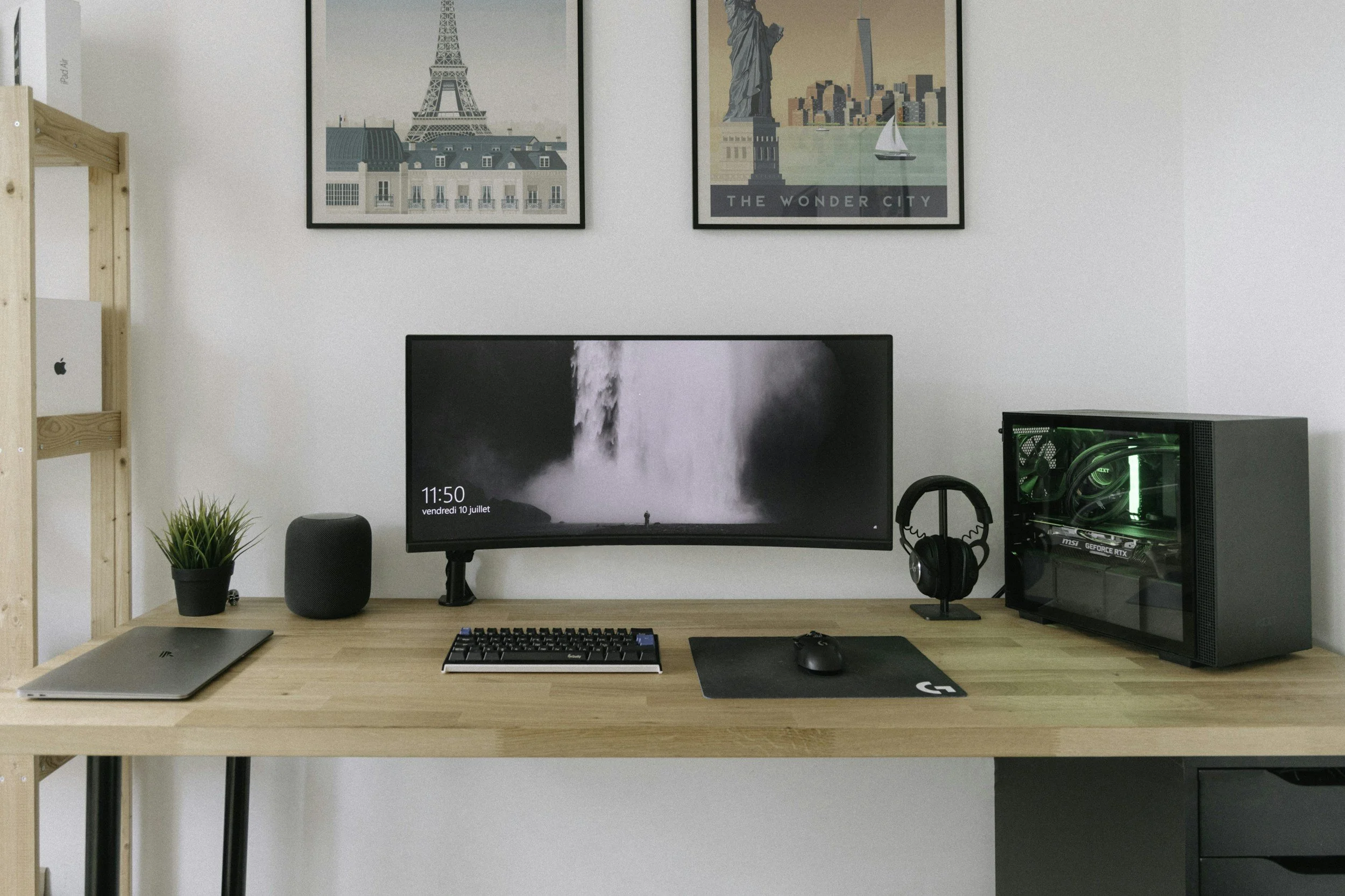 Modern computer desk setup with a curved monitor, desktop PC, keyboard, mouse, headphones, laptop, and decorative plant, with posters on the wall.