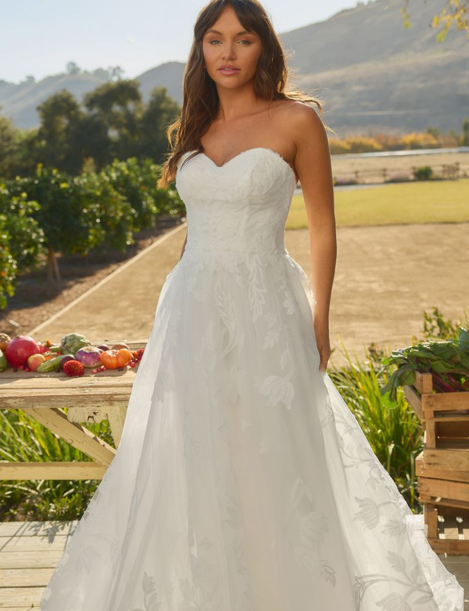 A woman in a white strapless wedding dress standing outdoors in a garden with a vineyard landscape in the background.