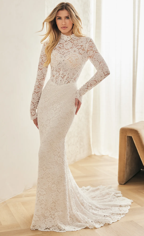 A woman in a long, elegant white lace wedding dress with long sleeves, standing indoors near a window with curtains, and a tan chair in the background.