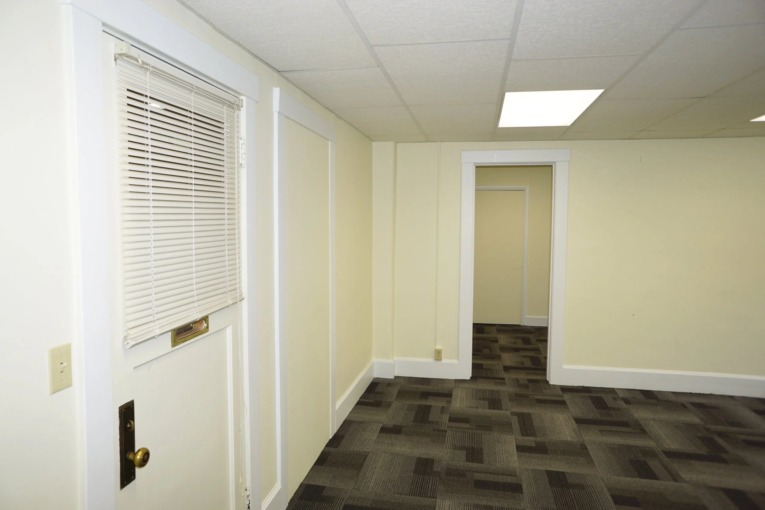 Interior of an empty office room with beige walls, a window with closed horizontal blinds, a door frame leading to another room, ceiling panels, and dark patterned carpet.