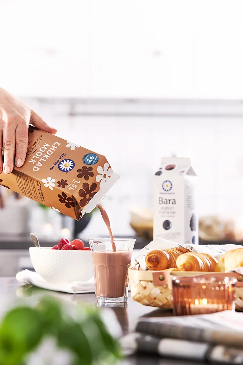 Person som häller chokladmjölk i ett glas på en köksbänk, omgiven av frallor, en skål med bär, ett paket yoghurt och ett ljus i ett glas.