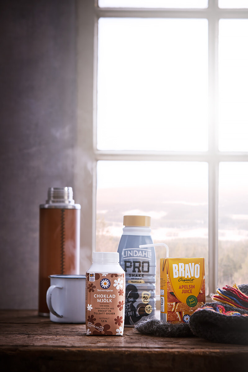 En bordstablett med en chokladmjölk, proteinshake, apelsinjuice, en kaffemugg, en termos, och ett par vantar framför ett fönster.