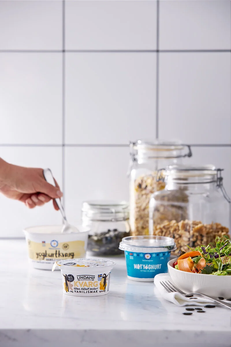 En hand som skedar yoghurt från en glasskål på ett vitt köksbord, bredvid olika typer av yoghurt i små burkar och stora glasburkar med müsli samt en sallad.