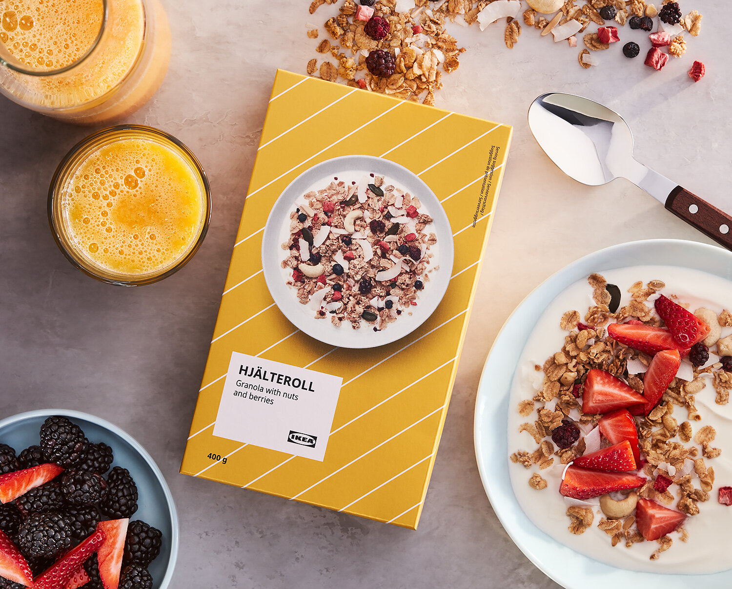 En bild av frukost med två glas apelsinjuice, en skål med jordgubbar och björnbär, en tallrik med yoghurt och granola toppad med jordgubbar, samt ett paket med granola med nötter och bär.