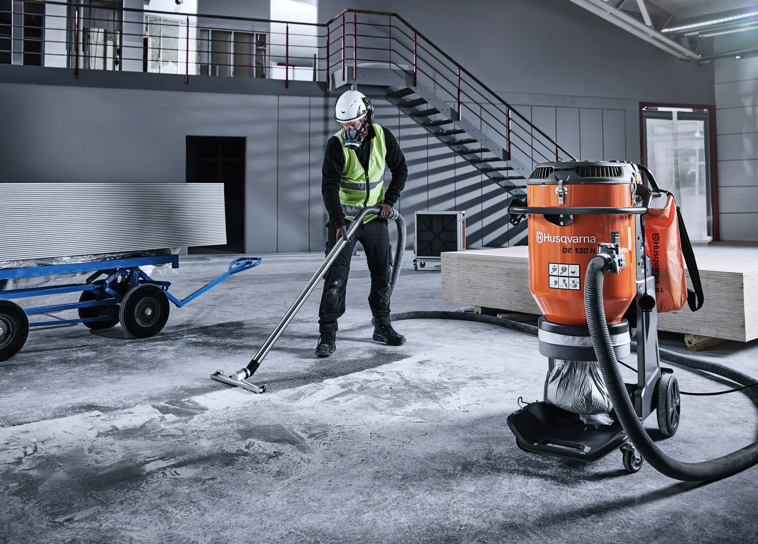 En person i skyddsutrustning arbetar med dammsugare inomhus. Rummet har metallräcken och en trappa i bakgrunden, samt byggmaterial och verktyg runt omkring.