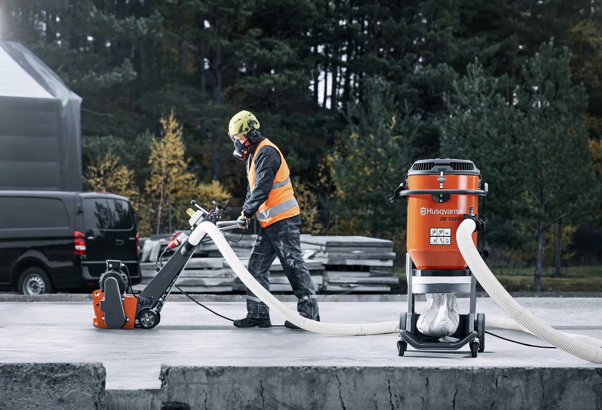 En person i säkerhetsutrustning använder en skogsavverkare i en skogsmiljö, med en orange Husqvarna dammsugare för skogsavverkning avfall i närheten.