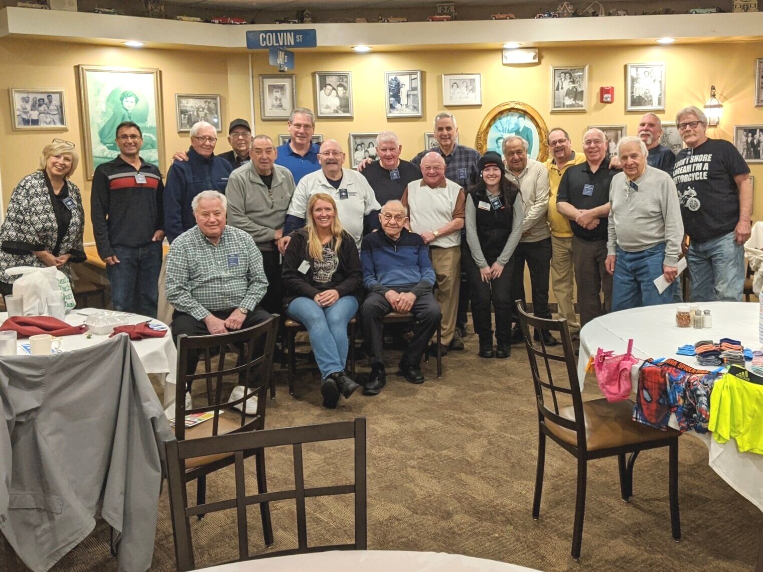 Group of people gathered in a restaurant for a photo, with some seated and others standing behind. The walls are decorated with photographs and a gold-framed mirror, with a blue street sign reading 'Colvin St' visible. Round tables with chairs and some table settings are in the foreground.